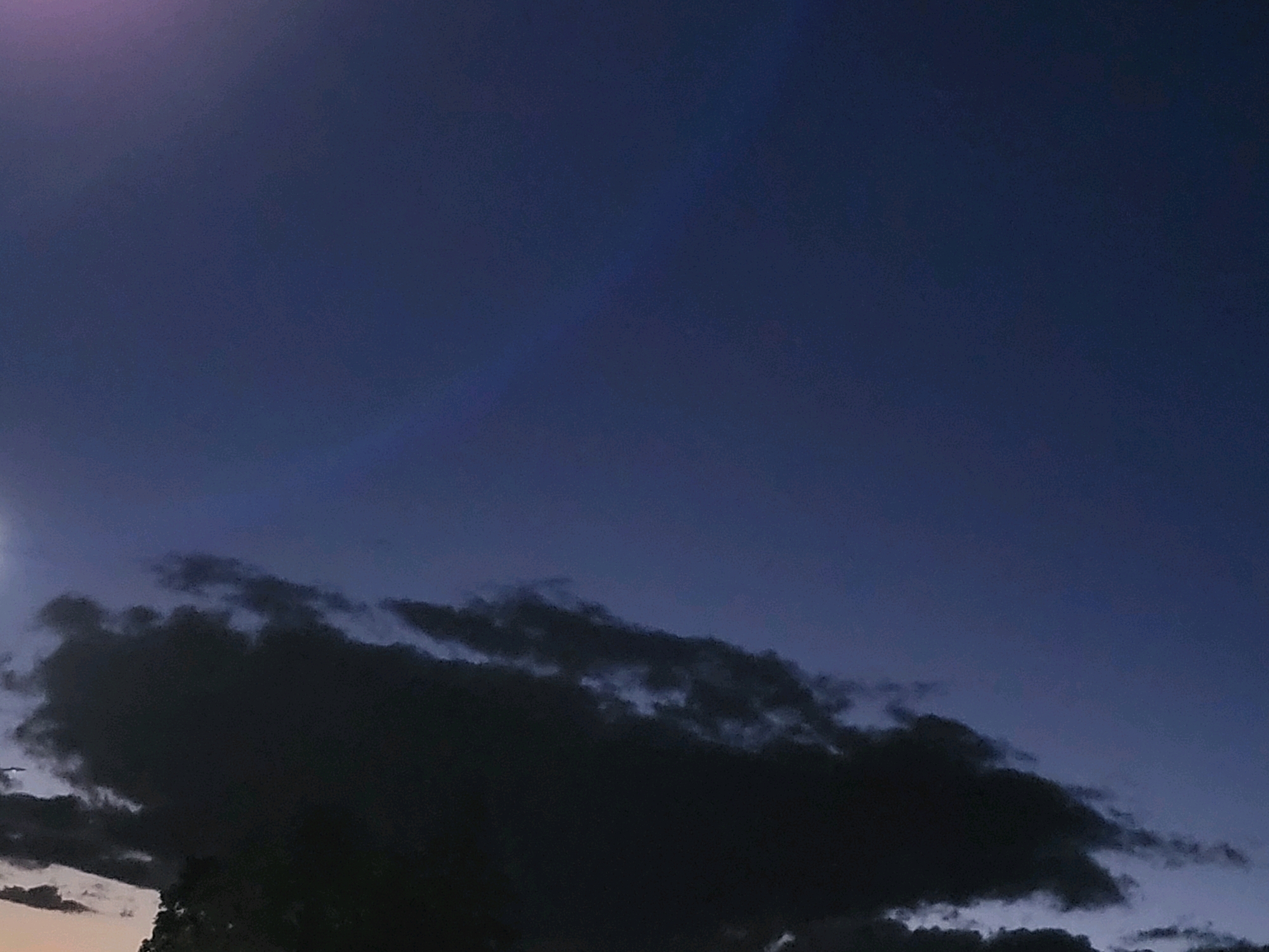 The image captures a dusky sky over Ozolnieki, Latvia. The upper portion of the frame is dominated by a deep blue, twilight sky, with a faint purple hue near the top left, possibly indicating residual sunlight or atmospheric conditions. A streak of light, possibly a lens flare, is visible in the upper right quadrant. The lower part of the image is filled with dark, brooding clouds that obscure the horizon. These clouds are dense and appear to be gathering, with some lighter patches suggesting breaks or thinner areas within the mass. The overall impression is one of an impending evening or a change in weather. No people or man-made structures are clearly identifiable in the image.
