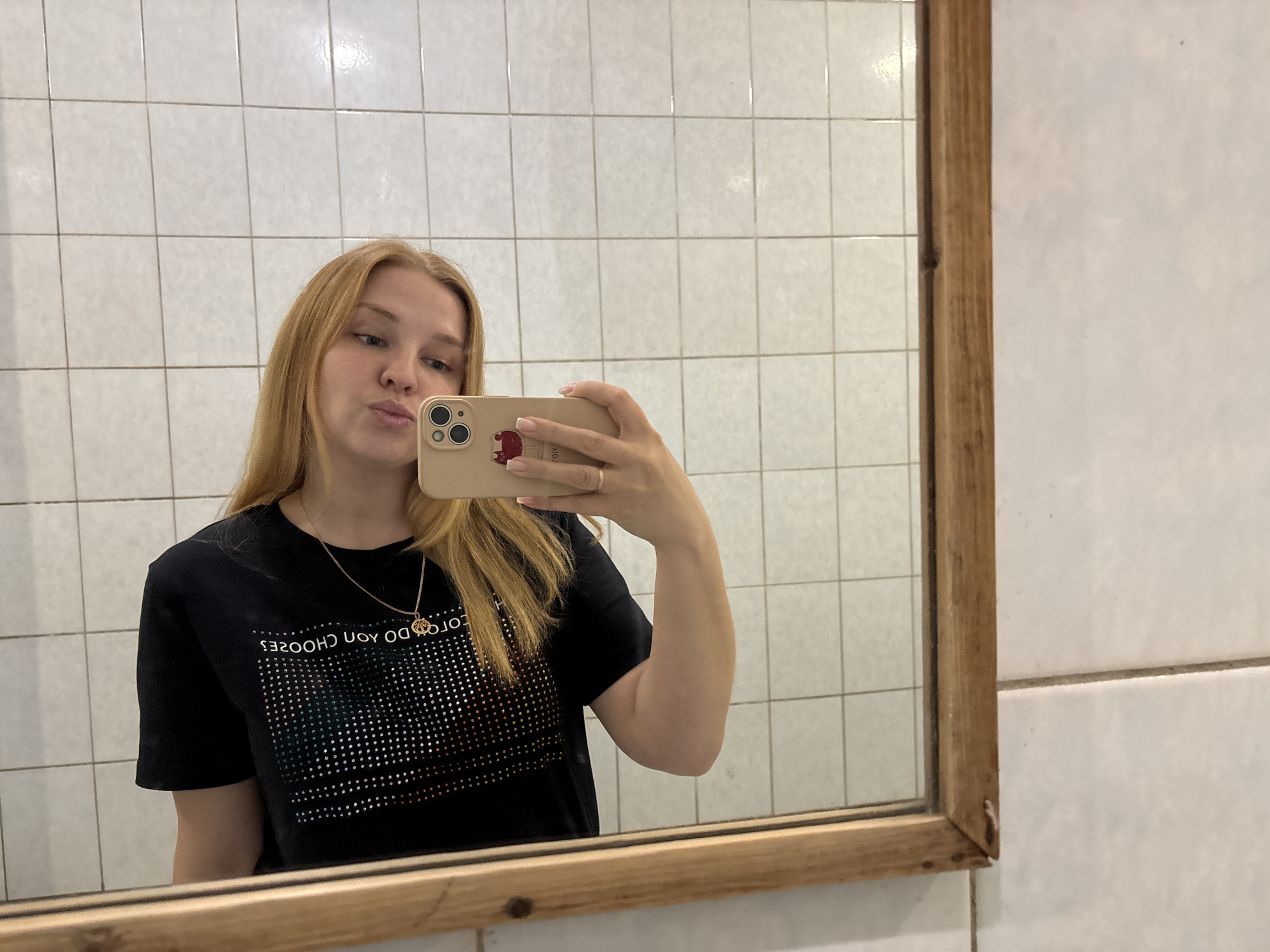 A young, blonde woman with fair skin is taking a selfie in a mirror. She is wearing a black t-shirt with a graphic design and a delicate gold necklace. Her phone has a beige case with a cartoon-like image on it, and she has a gold ring on her ring finger. The background consists of white square tiles, possibly in a bathroom. The mirror has a wooden frame that appears slightly aged. The location is Rusanovo, Russia.