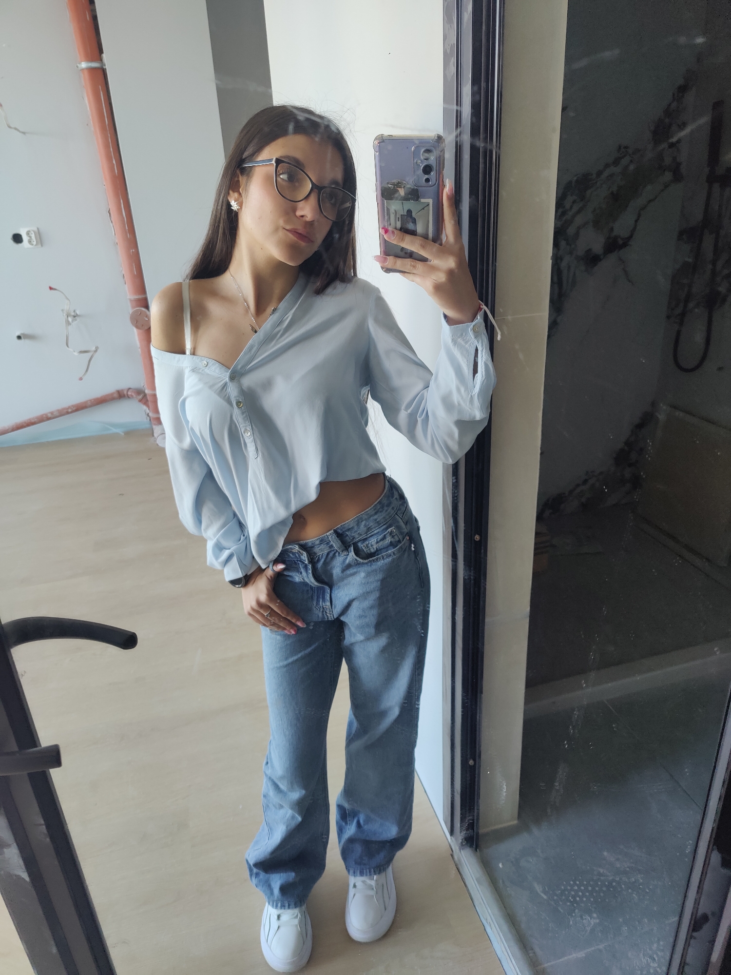 A young woman with long brown hair and glasses stands in front of a mirror, taking a selfie with her smartphone. She wears a light blue, button-down shirt, partially unbuttoned to reveal a bra strap, and blue jeans. She is wearing white sneakers. The background is a room under construction. In the mirror's reflection, there's an unfinished bathroom with gray tiling, and a plumbing fixture. The room also has exposed wiring and pipes. Although the precise location within Plovdiv, Bulgaria is unclear, the photo appears to be taken at an apartment under construction.