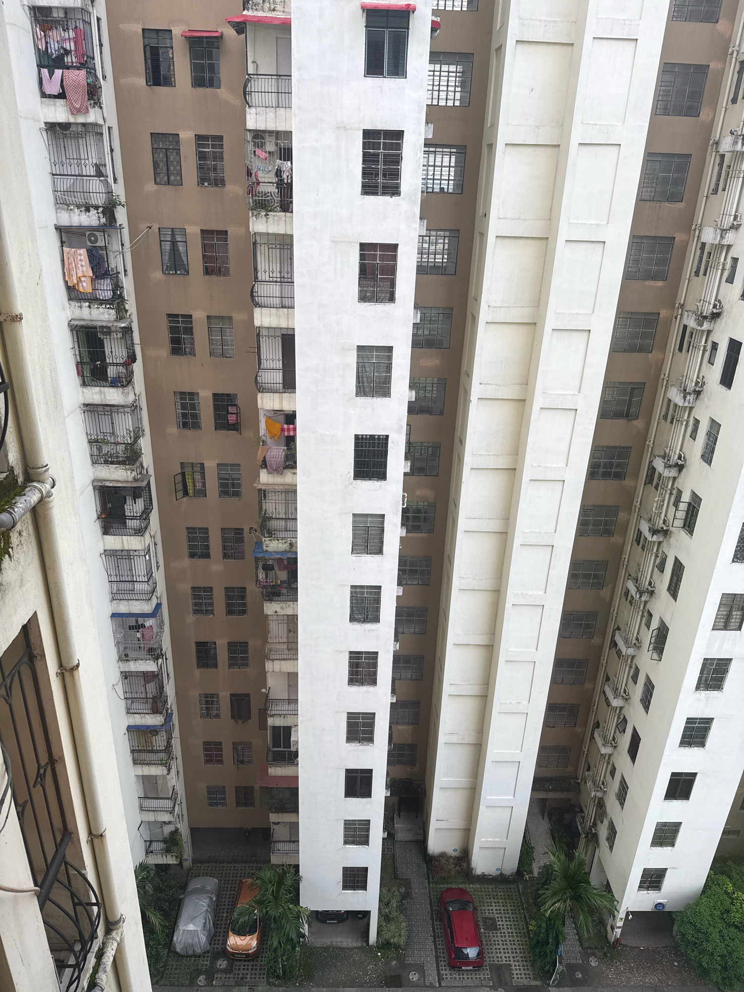 The image shows a view looking down on a dense urban environment in Kolkata, India. The scene is dominated by several tall, closely packed apartment buildings. The buildings are of various architectural styles and colors, with some appearing older and weathered, while others are more modern.
On the left side of the frame, a pale-colored building features balconies with laundry hanging out to dry. The windows have metal grates. Adjacent to this building is a taller structure with a light brown facade, also featuring balconies and windows with grates, and laundry visible. Further to the right, a white building with a more modern facade has rows of dark windows. Another building on the far right has a cream-colored exterior with visible pipes and ventilation systems running vertically along its side, and multiple windows, some with open shutters.
At the bottom of the image, the ground level is visible, showing a parking area with two cars: one silver car covered with a tarp and a red car parked. There are also some trees and greenery scattered around the parking area. The overall impression is one of a densely populated, multi-story residential area within a city.