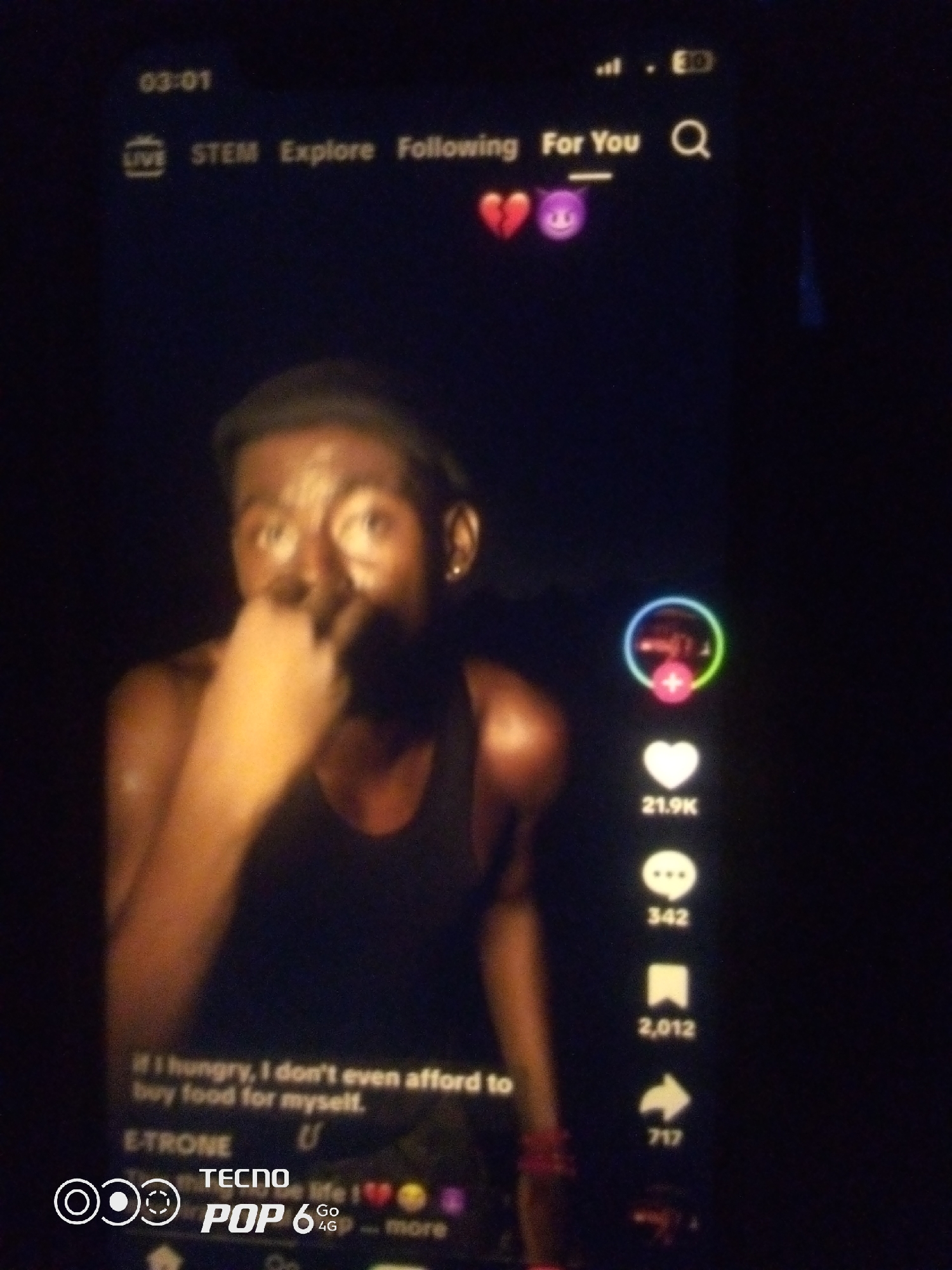 The image is a low-light, blurry, close-up photograph of a smartphone screen displaying a TikTok video. The video features a young man of African descent, with dark skin and short, dark hair, wearing a black tank top. He is looking at the camera with his hand near his mouth. The screen shows various TikTok interface elements, including a timer at the top left (03:01), navigation tabs like "LIVE," "STEM," "Explore," and "Following," and a "For You" section. There are also emojis, a broken heart and a devil face, near the "For You" tab. On the right side of the screen, there are icons for likes (21.9K), comments (342), shares (2,012), and an arrow for sharing (717). Text overlays at the bottom of the screen read "if I hungry, I don't even afford to buy food for myself." and "E-TRONE U". In the bottom left corner, there is a logo for "TECNO POP 6 4G". The background appears dark, suggesting the photo was taken in a dimly lit environment. The phone is likely being held by someone in Howayi, Nigeria, but this location is not explicitly visible in the image itself.