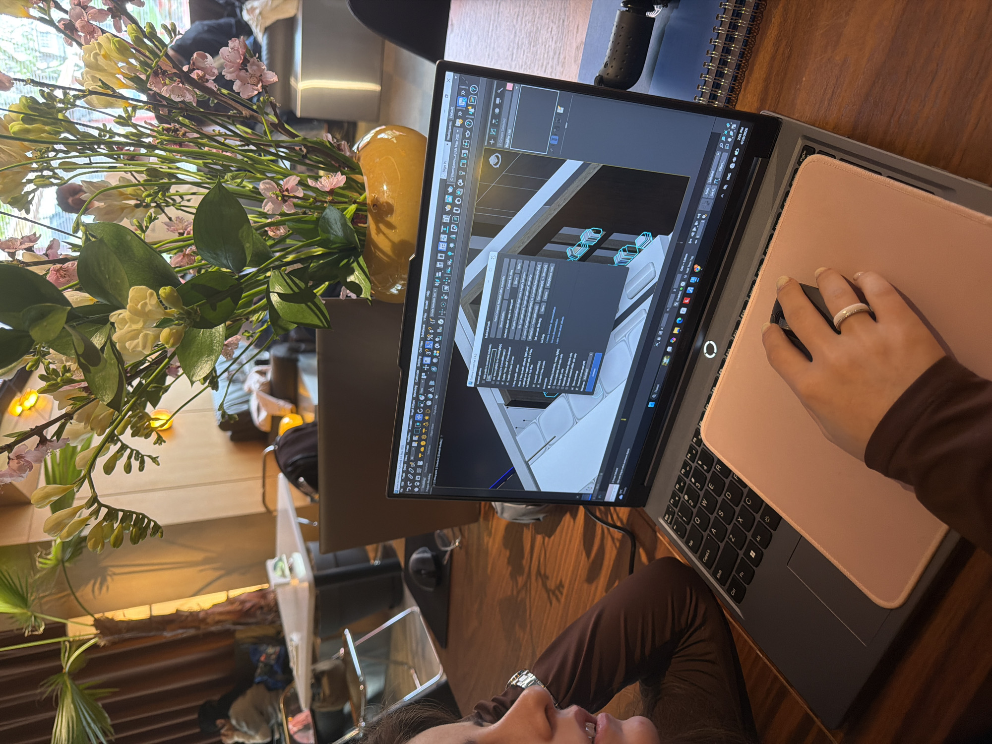 Here is a description of the image:
The image shows a person working on a laptop in what appears to be an indoor setting, possibly a cafe or workspace. The laptop screen displays a 3D modeling or architectural software, suggesting the person is engaged in design or drafting work. A pink mousepad sits on the keyboard area of the laptop, with the person's hand visible operating a mouse. The individual is wearing a brown long-sleeved garment, and a ring is visible on their finger.
A vase of flowers, featuring a mix of white and pink blossoms, is placed near the laptop. The table surface is wooden. The lighting suggests a natural or well-lit environment.
The location is possibly in Yerevan, Armenia, based on the language displayed on the laptop.