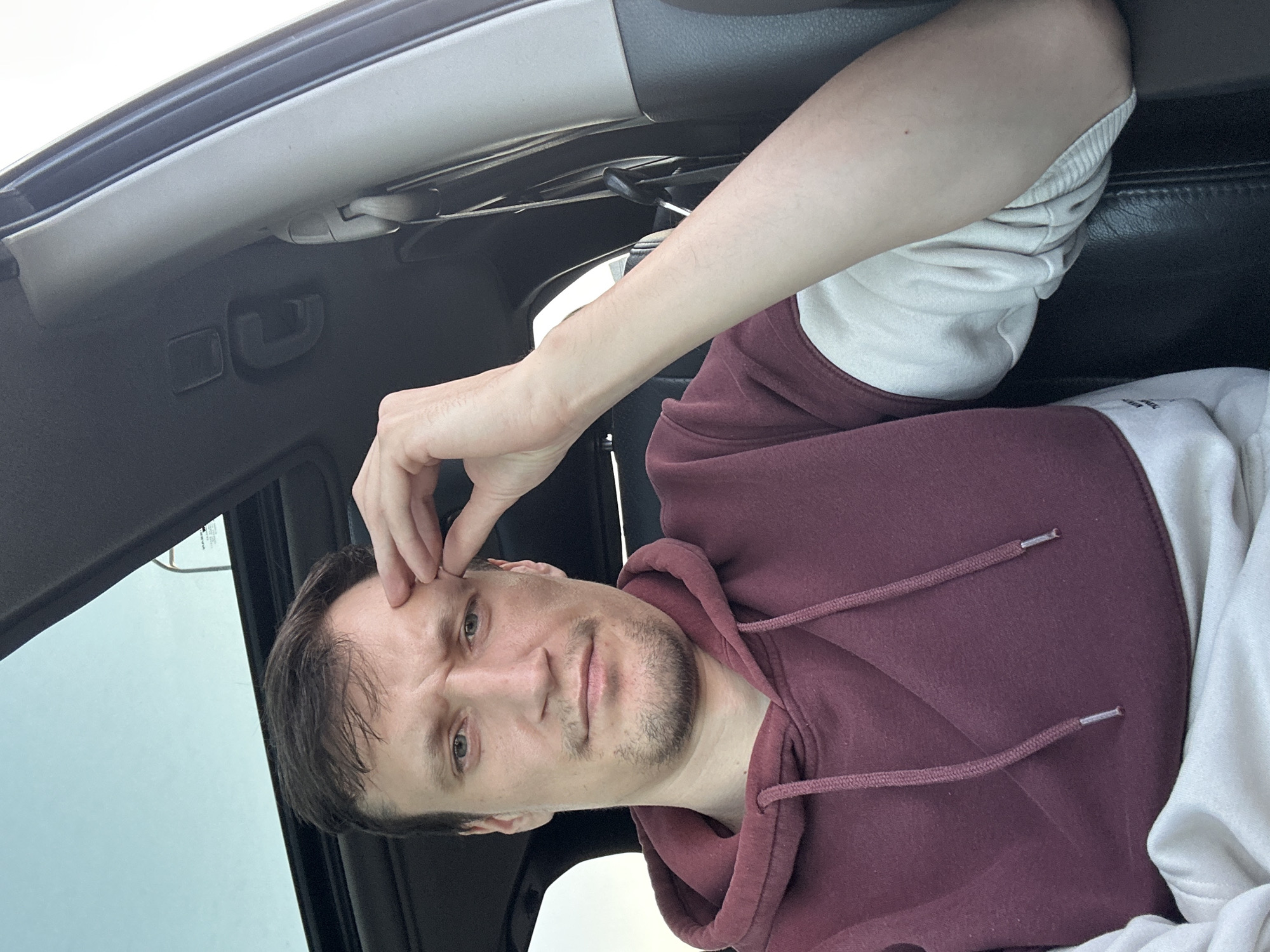 A man is sitting inside a car, holding his forehead with his right hand. He is wearing a maroon hoodie with cream-colored sleeves and is looking towards the camera with a concerned expression. The car's sunroof is open, revealing a cloudy sky. The interior of the car appears to be dark, with black leather seats and gray trim. There are no identifiable landmarks or signs that would indicate the specific location within Kamianets-Podilskyi, Ukraine.