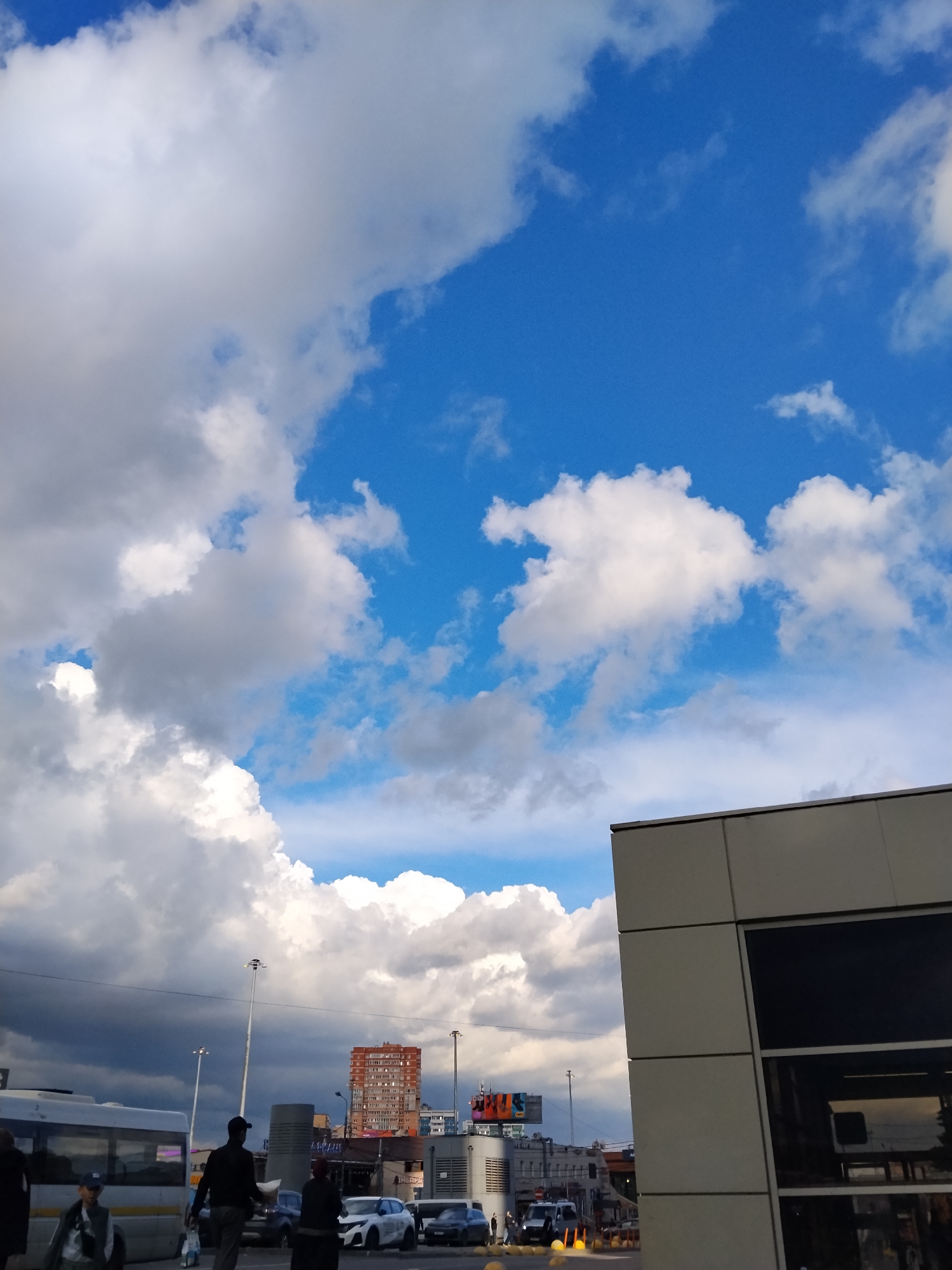 The image shows an urban scene in Kotelniki, Russia. A partly cloudy sky fills the upper portion of the frame, transitioning from bright blue to greyish-white cumulus clouds. In the lower half, street-level activity is visible. A bus is parked on the left side. Pedestrians walk along the sidewalk. A red brick high-rise building is visible further in the background, along with other urban structures, streetlights, and billboards. Cars are parked along the street. The right side of the frame is dominated by a modern building with a light-colored exterior.