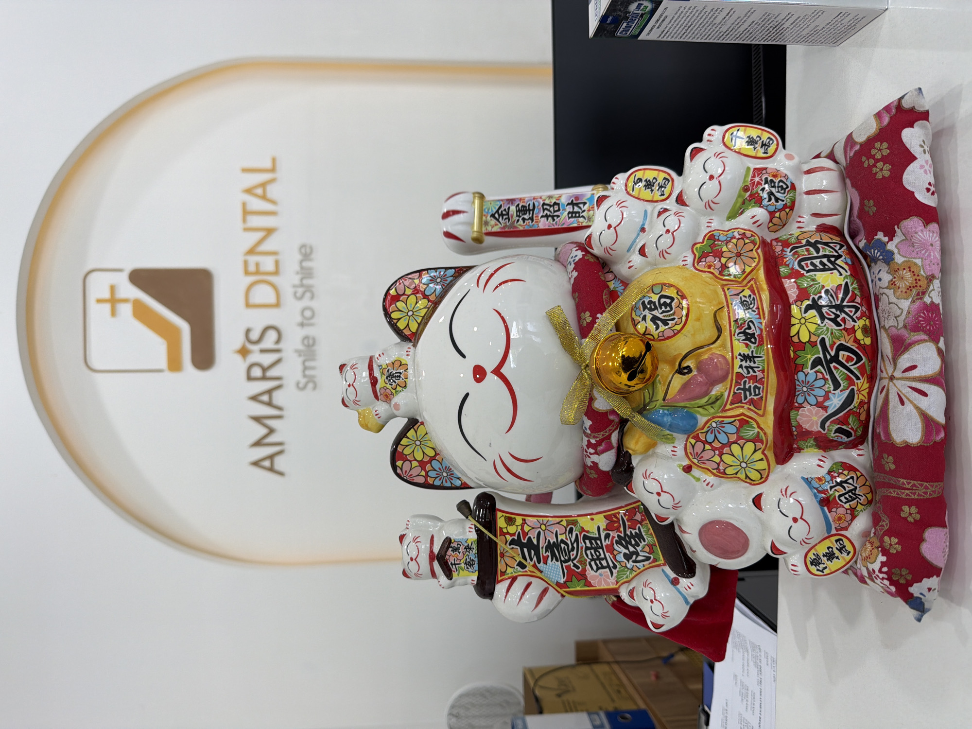 This indoor photograph, taken in a dental clinic in Đà Nẵng, Vietnam, features a large, ornate Maneki-neko (beckoning cat) figurine prominently displayed. The Maneki-neko is predominantly white with red, gold, and colorful floral patterns, and its left paw is raised in a beckoning gesture. Several smaller Maneki-neko figures are integrated into its design, perched on the larger cat and holding golden coins or scrolls. The main cat also holds a large gold coin and a decorative banner. Behind the figurine is a red cushion or piece of fabric adorned with white and pink floral patterns.
In the background, a white wall displays the logo and name of "AMARIS DENTAL" with the tagline "Smile to Shine." The logo features a brown and gold abstract design incorporating a plus sign. To the right of the Maneki-neko, a black object, possibly a computer monitor or part of a shelf, is visible, with a white box labeled "Chlorhexil" on top. Below the Maneki-neko, glimpses of papers or documents are visible.
The scene suggests a welcoming commercial environment, likely the reception area of the dental clinic, where the Maneki-neko serves as a symbol of good luck and prosperity for the business.
Visible text includes:
- On the wall: "AMARIS DENTAL", "Smile to Shine".
- On the Maneki-neko figures and banners (in Chinese/Japanese characters): "金運招財" (attracting wealth and good fortune), "千萬両" (ten million gold coins, appearing twice), "百萬両" (one million gold coins), "億萬両" (one hundred million gold coins), "福" (fortune/blessing, appearing multiple times), "吉祥如意" (good fortune as desired), "客來" (customers come), and "隆" (prosperous).
- On a box in the upper right: "Chlorhexil".