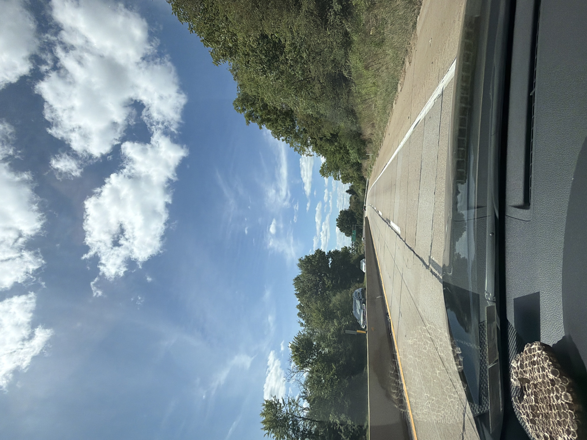 The image shows a view from inside a vehicle traveling on a highway in Findlay Township, United States of America. The road is paved with concrete and marked with white lines. The vehicle is traveling in the right lane. On the left side of the road, there is a concrete barrier and trees. On the right side of the road, there are trees and grass. The sky is blue with white clouds. There are other vehicles on the road. In the foreground, on the dashboard, there is a decorative object that appears to be made of a honeycomb-like material.
