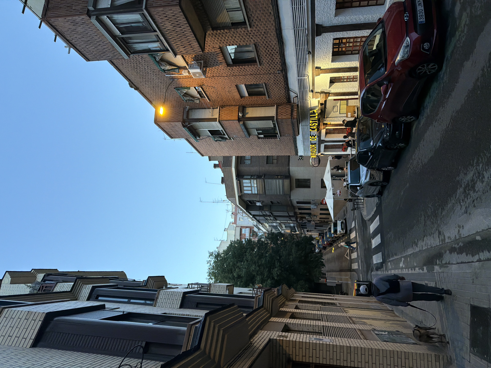 The image depicts a street scene in Madrid, Spain. The photo is oriented vertically, showing the street running in the distance. Buildings flank both sides of the street, varying in architectural styles with brick facades and multi-story structures. A tree with full foliage stands mid-street. Cars are parked on the right side of the street, including a red car at the top right of the frame. People are visible, including a person walking a dog on the sidewalk at the bottom right, and several individuals at outdoor seating near a business marked "MIRADOR DE CASTILLA". The sky is a clear blue.