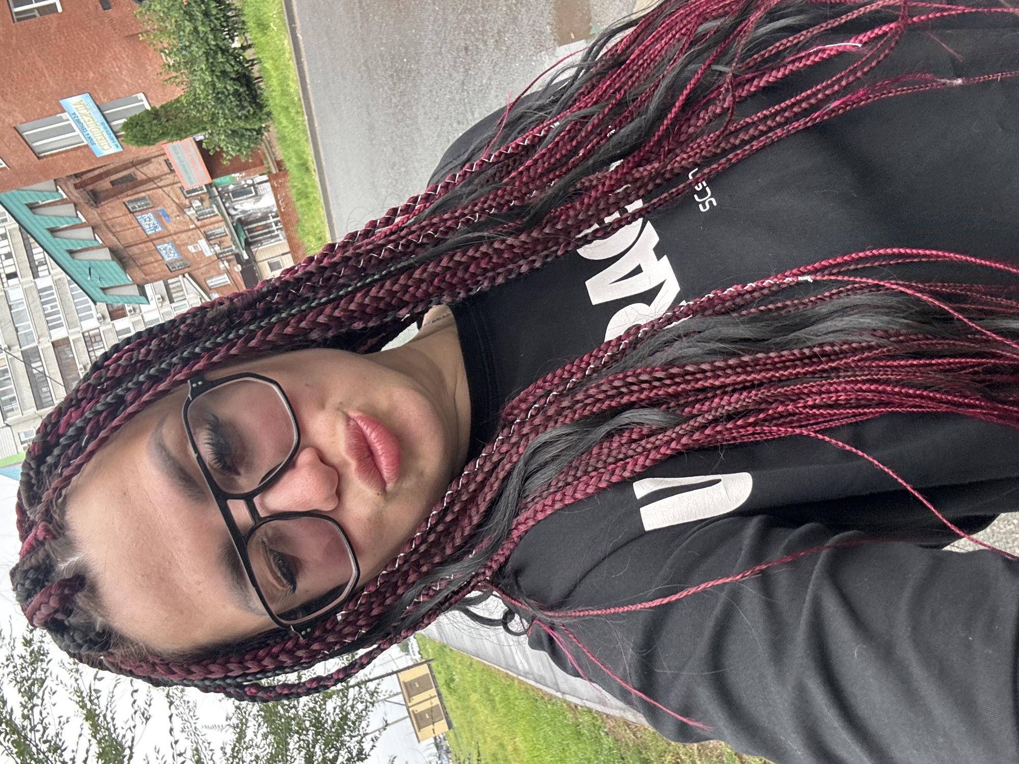 A woman with long, dark, and maroon braided hair, wearing large black-rimmed glasses and a black sweatshirt with white lettering, poses for a selfie outdoors. The background features a street, a grassy area, and multi-story buildings. The woman is facing the camera with her lips pursed. The scene appears to be on a cloudy day. The image is identified as being taken in Tolyatti, Russia.