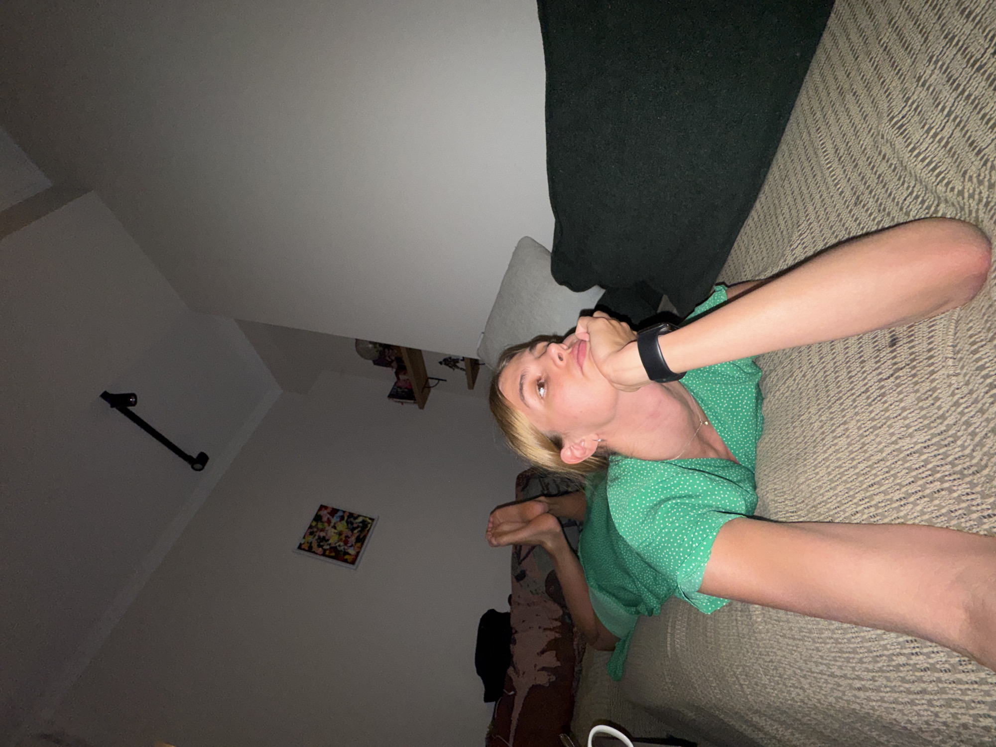 A young person with blonde hair, wearing a green polka-dot dress with white spots, lies on a textured, light-brown sofa in a dimly lit indoor setting. The image is taken from a low, angled perspective, looking up towards the ceiling and walls. The person's right hand is resting near their mouth or chin, and a dark smartwatch is visible on their left wrist. Their feet are curled up. A dark blanket or throw is draped over part of the sofa. In the background, a white wall features a small, square, colorful framed picture with an abstract or pixelated design, and a black wall-mounted track light fixture with two spotlights. The overall lighting suggests evening or night, with soft artificial light illuminating parts of the room. The person's expression appears relaxed or contemplative. No text is visible.