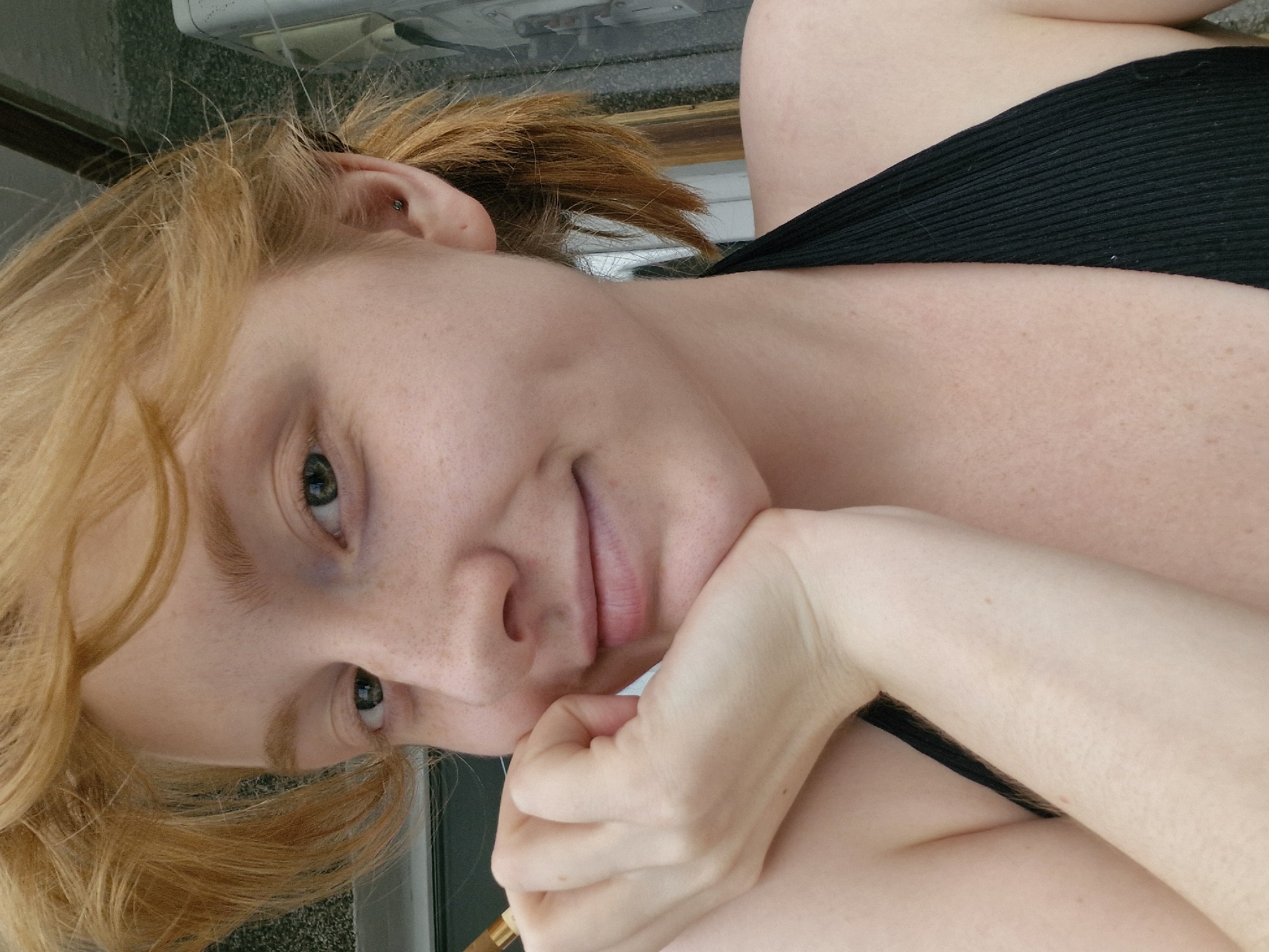 A young woman with fair skin, freckles, and reddish-blonde hair is shown in a close-up, self-taken photograph. She is wearing a black halter-neck top and rests her chin on her hand, with her elbow on her arm. Her eyes are looking towards the camera, and she has a slight smile. The background appears to be an outdoor setting with hints of a building structure. The image does not contain enough information to determine its specific location in Yekaterinburg, Russia.