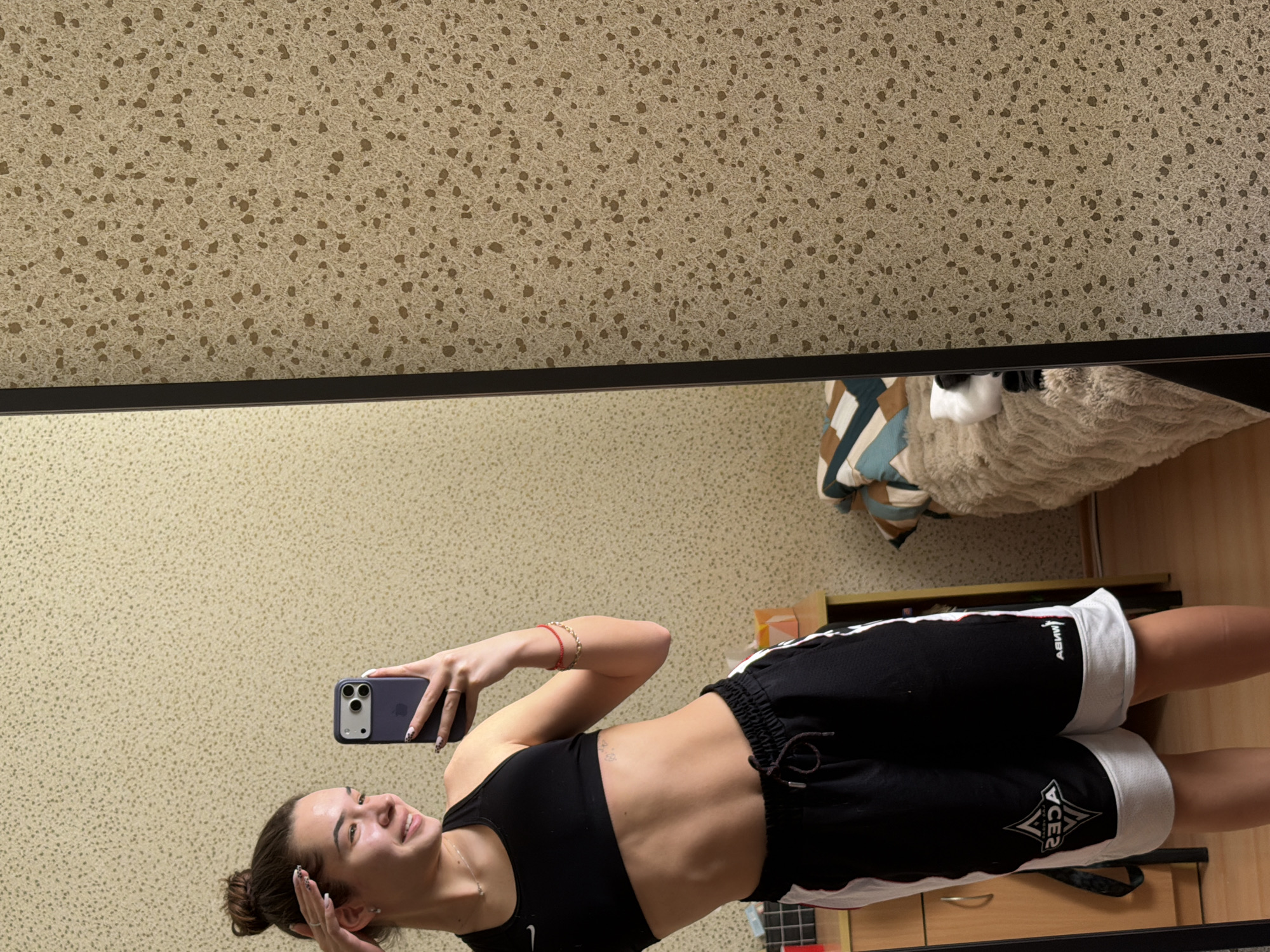 A female stands in front of a mirror, holding a cell phone in her right hand, likely taking a selfie. She is wearing a black sports bra and black shorts with white trim. The room's wallpaper has a textured, speckled pattern in beige and brown. The mirror's frame is black. Visible behind the woman and to the right of the mirror is a wooden cabinet, part of a couch with patterned pillows, and a textured, cream-colored blanket or cushion. The location is likely an interior space, potentially a bedroom or dressing area. Given the context of previous requests, the implied location is Sofia, Bulgaria.