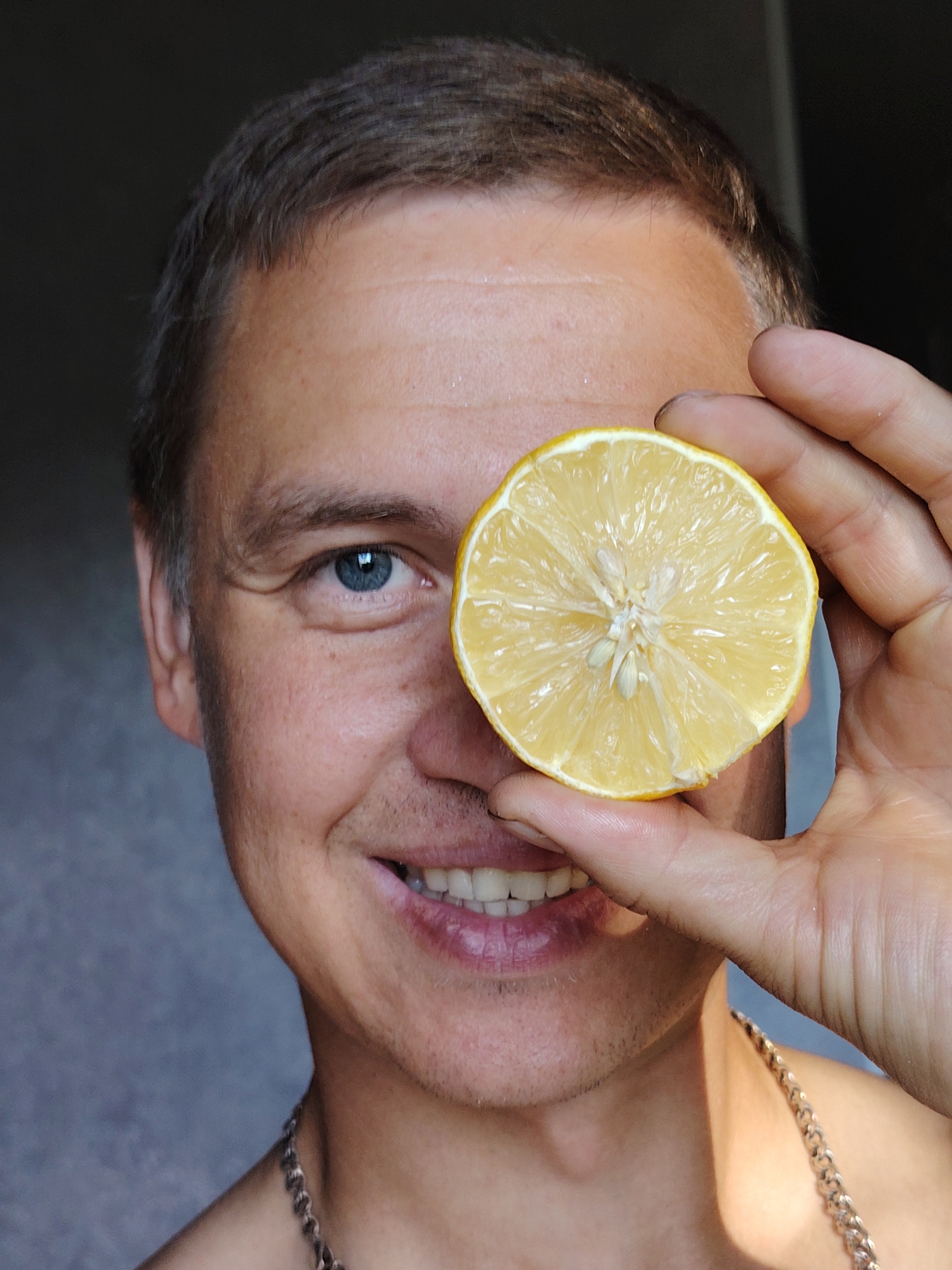 The image is a close-up shot of a man's face. He is holding a slice of lemon over his right eye, obscuring it. His left eye, which is visible, is blue. He has short, brown hair and is smiling, showing his teeth. He is wearing a silver chain around his neck. The background is a plain, greyish wall. The lighting suggests the photo was taken indoors, possibly near a window, with sunlight casting a shadow on the right side of his face. There is no identifiable location within the city of Chaikovsky, Russia.