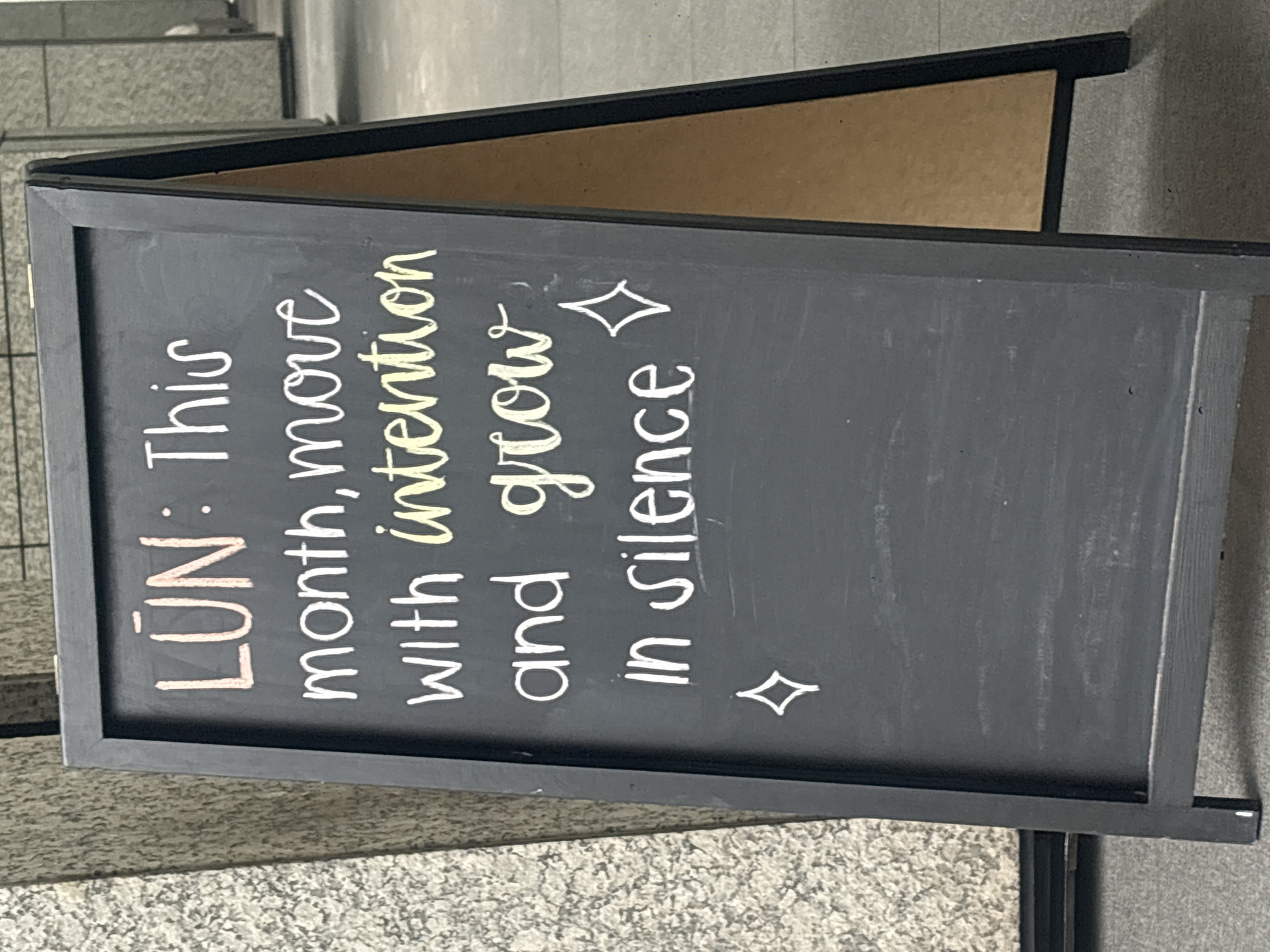 A black chalkboard sign is resting on a gray tile surface. The sign reads "LUN: This month, move with intention and grow in silence," with "intention" colored in yellow and diamond shapes appearing at the beginning and end. The back of the sign is a brown color. The photo was taken in Mexico City, Mexico.