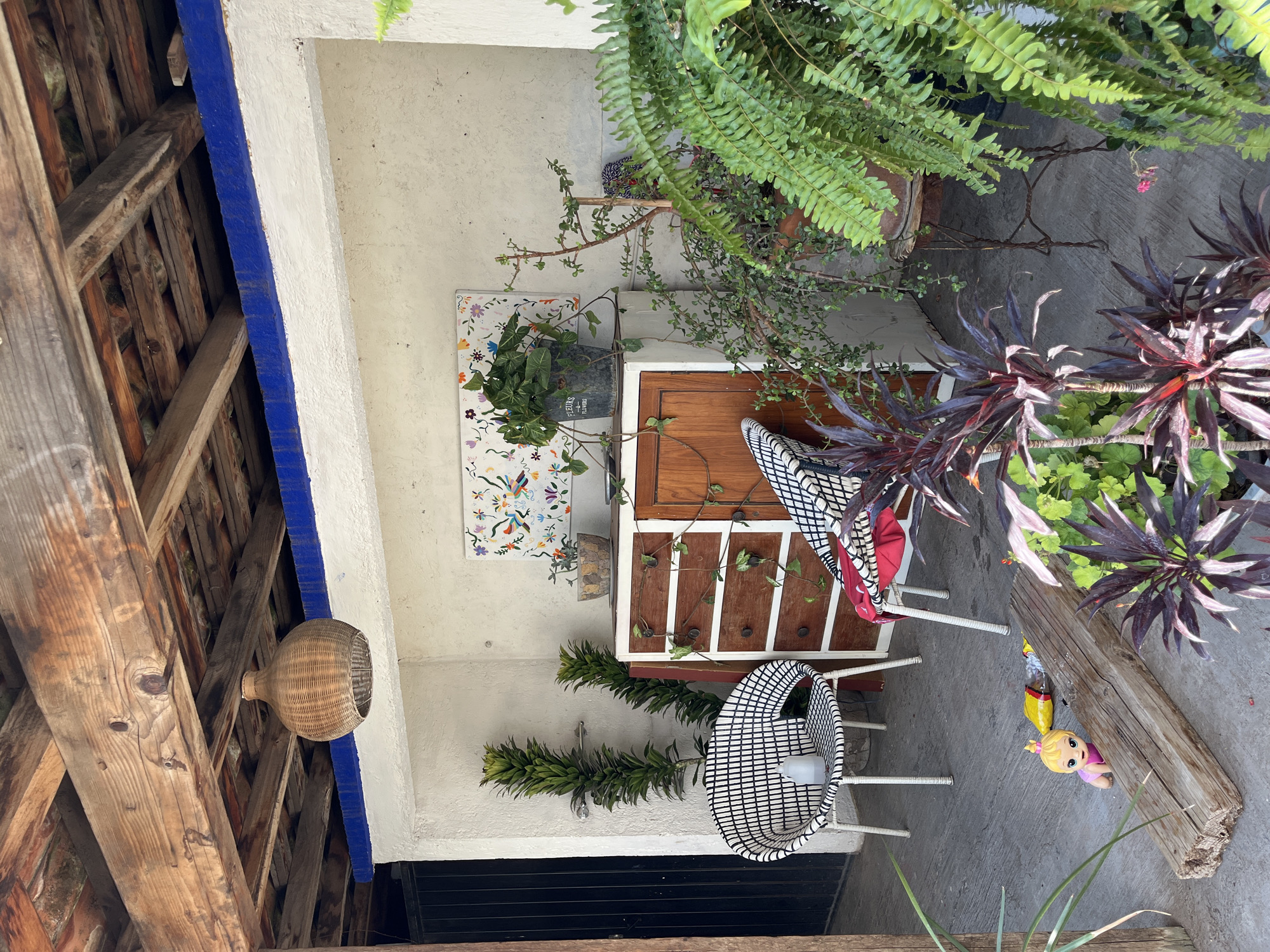 The image depicts an outdoor patio in what is likely Puebla, Mexico, given the architectural style and decor. The patio is enclosed by white walls and a wooden beam roof structure, painted with blue trim.
A wooden dresser with white trim stands against the wall, adorned with potted plants. Decorative tiles hang above the dresser. Two woven chairs with white metal legs are placed in front of the dresser.
Several potted plants with varied foliage provide lush greenery to the scene. A woven, gourd-shaped object hangs from the wooden beams above. A doll rests on the ground near a wooden beam. The patio floor appears to be made of concrete. The image is taken from a high vantage point.