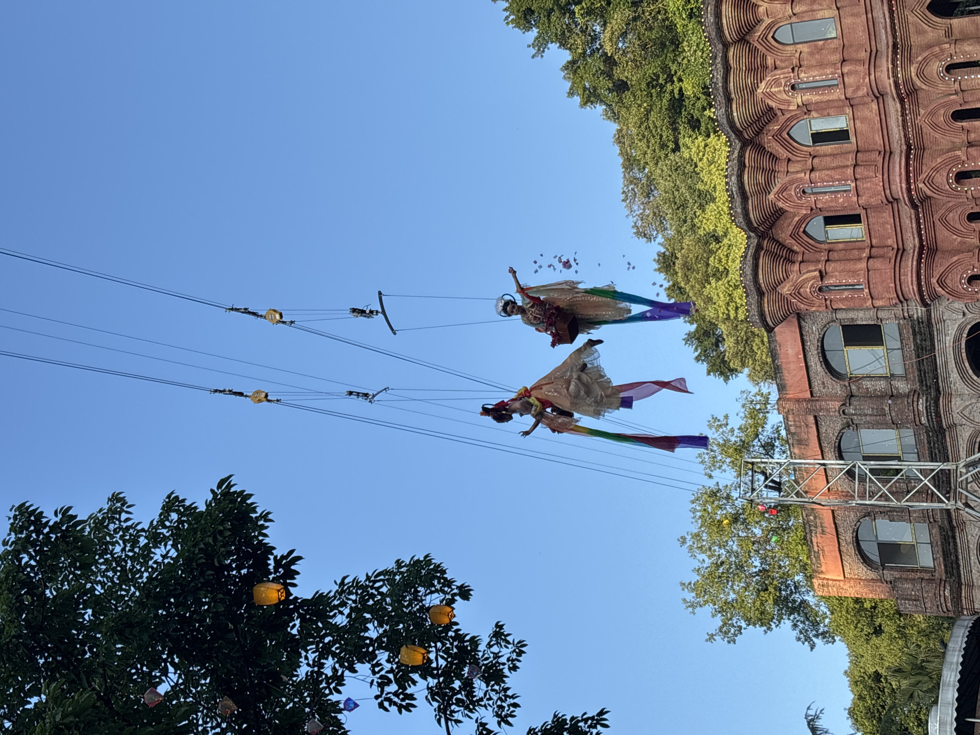 Two performers in elaborate costumes are suspended in the air by wires against a clear blue sky. The performer on the left is wearing a flowing, light-colored dress and a headdress, with rainbow-colored fabric streamers attached to her. The performer on the right also wears a similar dress and headdress, holding a basket from which petals are falling. Both performers appear to be part of an aerial performance. Below them, a distinctive red brick building with arched windows and ornate detailing is visible, suggesting it might be a historical or cultural landmark. Green trees line the background, and a metal structure, possibly for supporting the aerial performance, is also present. The scene is lit by natural sunlight, creating a bright and clear atmosphere. The image was taken in the city of Baqiao, China.