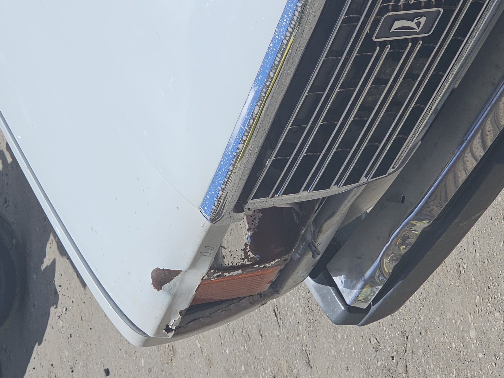 The image shows a close-up, angled view of the front corner of a white car. The car appears to be damaged, with rust and chipped paint visible on the fender and around the headlight area. A portion of the car's grille, which is silver with vertical slats and a dark emblem in the center, is also visible. The grille is bordered by a chrome trim piece that is also showing signs of wear. A reflection of the surroundings can be seen on the chrome bumper below the grille. The car is parked on a paved surface, likely a road or parking lot, with some small stones and debris visible. The lighting suggests it is daytime, and the shadows indicate a sunny environment. The overall impression is that of a vehicle that has been in an accident or is in a state of disrepair. No people are visible in the image. No text is legible in the image. The context provided indicates the location is Nizhny Novgorod, Russia.