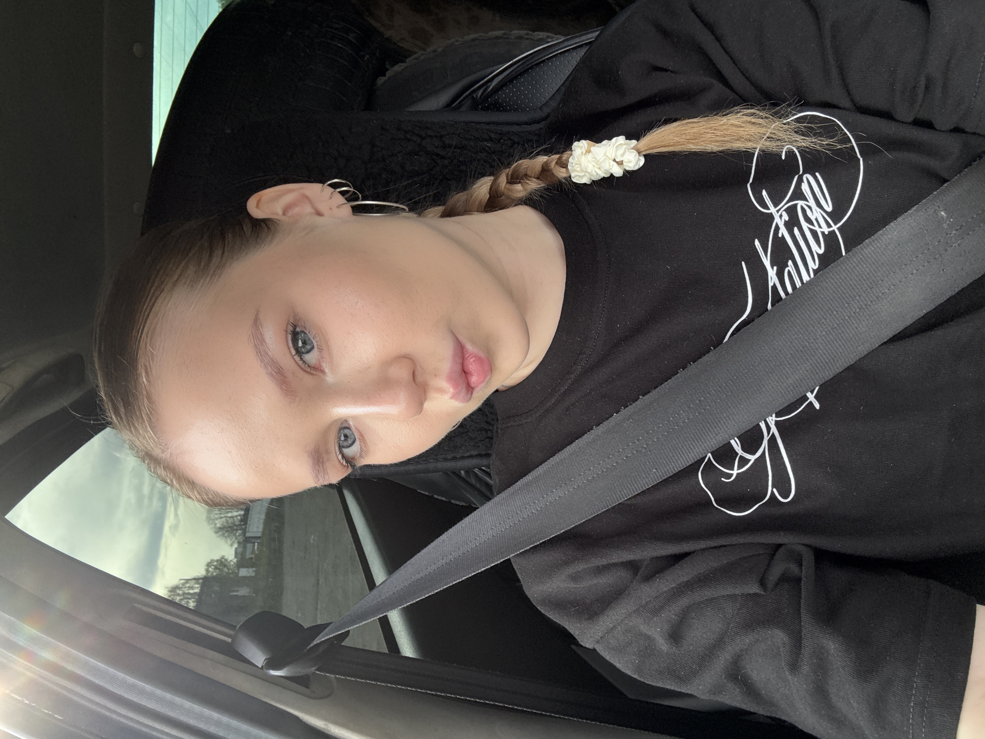 A young woman is in the driver's seat of a car, wearing a black t-shirt and a seatbelt. Her blonde hair is braided, with a white hair tie at the end of the braid. She is looking at the camera with a slight pout. Outside the car window, trees and buildings are visible under a cloudy sky. The location is Chaikovsky, Russia.