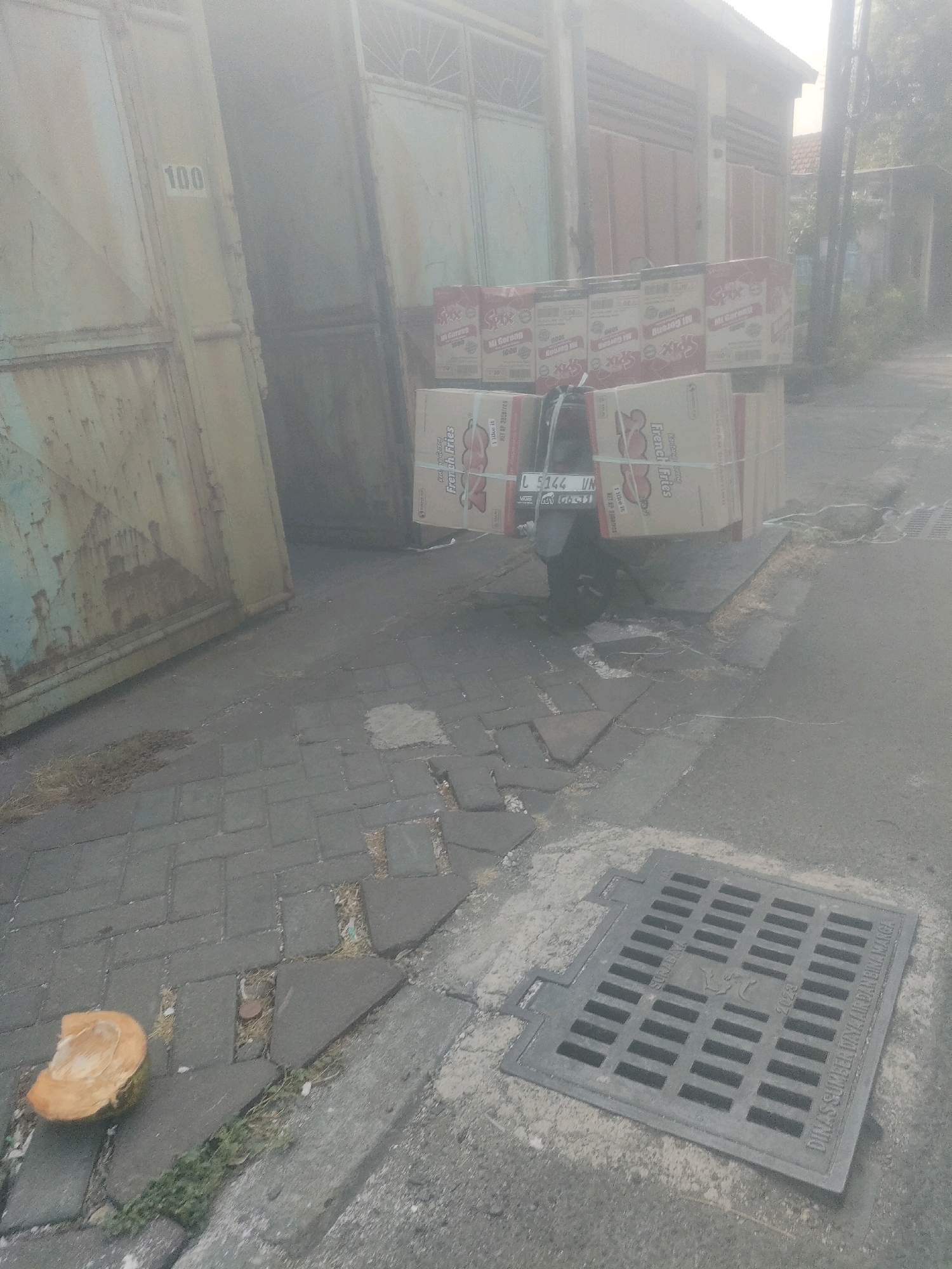 In the city of Surabaya, Indonesia, a motorbike is parked on a paved area next to a large, weathered metal gate with the number "100" stenciled on it. The motorbike is loaded with stacked cardboard boxes containing products. Several boxes are labeled "Mi Goreng" and "French Fries". A damaged, half-eaten coconut lies on the paved ground to the left of the motorbike. To the right of the motorbike, a large metal grate covers a drainage opening. The scene appears to be outdoors, possibly in an alley or on a street with a building in the background. The lighting suggests it is daytime, but the image is hazy, possibly due to dust or fog. There are no people visible in the image.