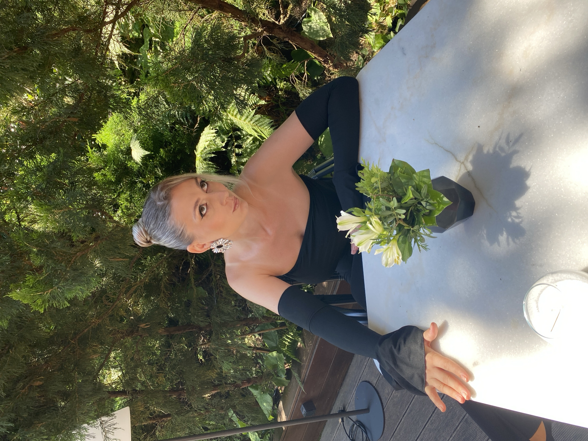 A woman with her hair in a bun and large sparkly earrings sits at a table outdoors. She wears a strapless black dress with long black sleeves that extend to her wrists. Her left arm rests on the table, and her left hand is splayed on the surface. A small vase with white flowers and greenery sits on the table in front of her. The background features lush green foliage and trees. The scene appears to be an outdoor seating area, possibly a cafe or restaurant, set within a garden or park environment, consistent with locations found in Sochi, Russia.