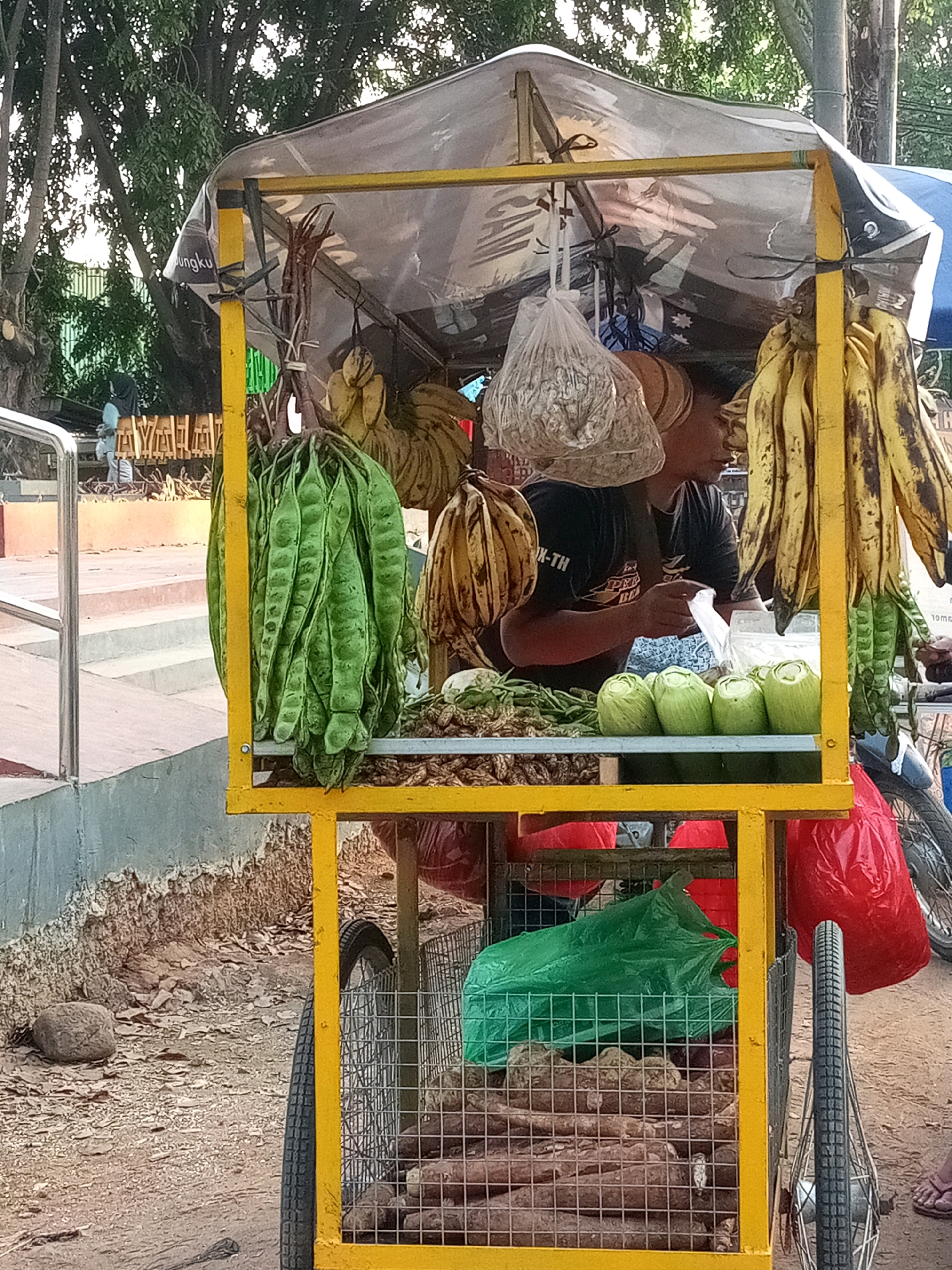 This image depicts a street vendor's cart in Bekasi, Indonesia, laden with fresh produce. The cart, painted bright yellow, is covered by a translucent plastic tarp, providing shade. Hanging from the cart are bunches of bananas and a large cluster of green, pod-like vegetables, likely petai (stink beans). In the foreground, several rounded, pale green vegetables are neatly arranged, possibly gourds or young coconuts. Behind them, a vendor, wearing a dark t-shirt, is visible from the chest up, handling a plastic bag. Below the main display, the cart's lower section is a wire cage containing more root vegetables, covered by green and red plastic bags. The scene is set on a dirt and gravel path, with a concrete barrier and stairs to the left, suggesting a public space or roadside. The overall impression is a typical scene of local commerce in an urban Indonesian setting.