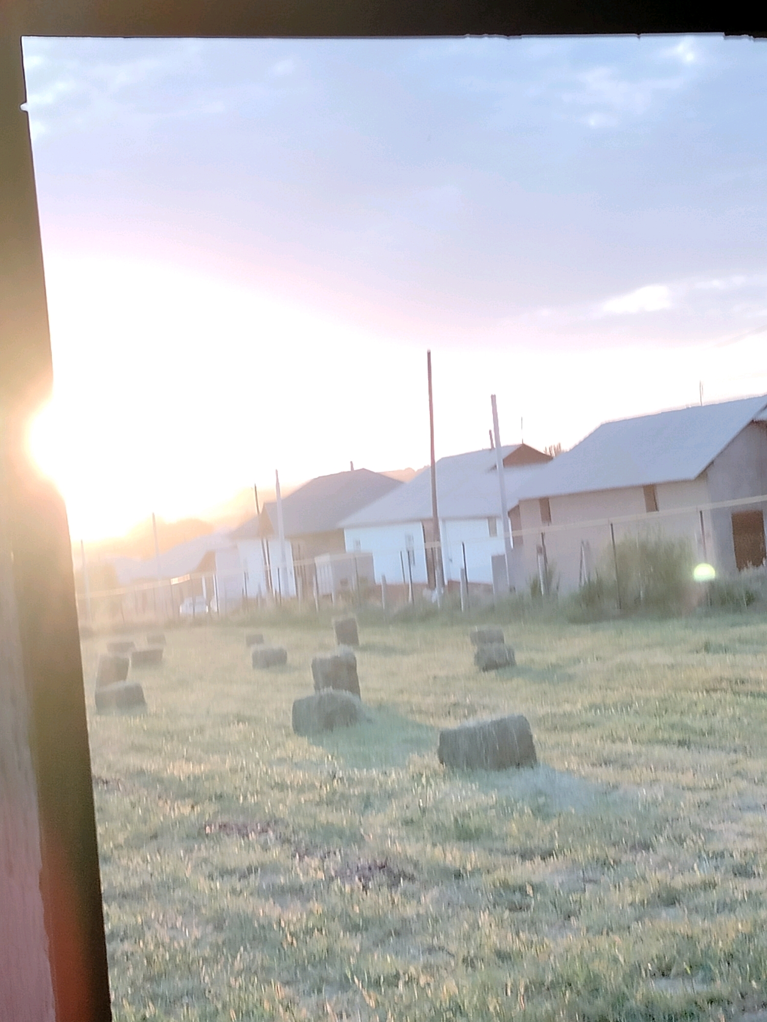 The image captures a rural landscape at sunrise, characterized by a bright sun in the upper left quadrant, casting a warm glow across the scene. In the foreground, a field is dotted with numerous hay bales. Beyond the field, a row of houses with gray roofs is visible, indicative of a residential area. The sky is a soft gradient of blue and pink, suggesting early morning light. This scene is located in the city of Kölkent, Kazakhstan.