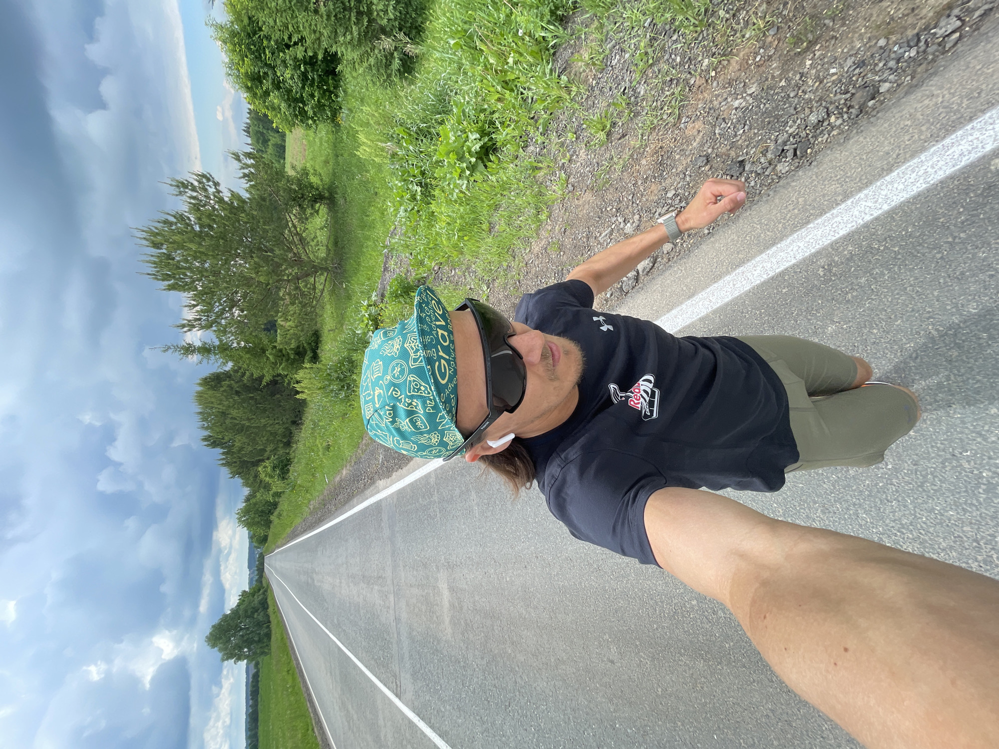A male individual is captured in a dynamic selfie on a long, open road, extending his right arm to hold the camera. He is seen from a low angle, seemingly leaning or in motion, with the road stretching into the distance behind him.
The subject is wearing a light teal-colored cap with various patterns and words like "Grave," "Pizza," and "Adventure" visible. He sports dark sunglasses and a black t-shirt with a red and white logo on the left chest, partially obscured, which includes the word "Red." White wireless earphones are visible in his ears. His right arm, holding the camera, has a silver-banded watch on the wrist. He wears olive green shorts or pants, and his footwear is mostly out of frame, though a sliver of a white shoe is visible.
The scene is an outdoor setting on an asphalt road with prominent white lines marking the side. To the left of the road, beyond a gravel shoulder, are green fields and scattered trees. The horizon shows distant hills or land formations. The sky above is largely cloudy, with patches of lighter blue visible. The overall impression is of a bright, possibly overcast day in a rural or less populated area. The person's facial expression, though largely hidden by sunglasses, suggests a relaxed and possibly playful mood given the dynamic pose.
