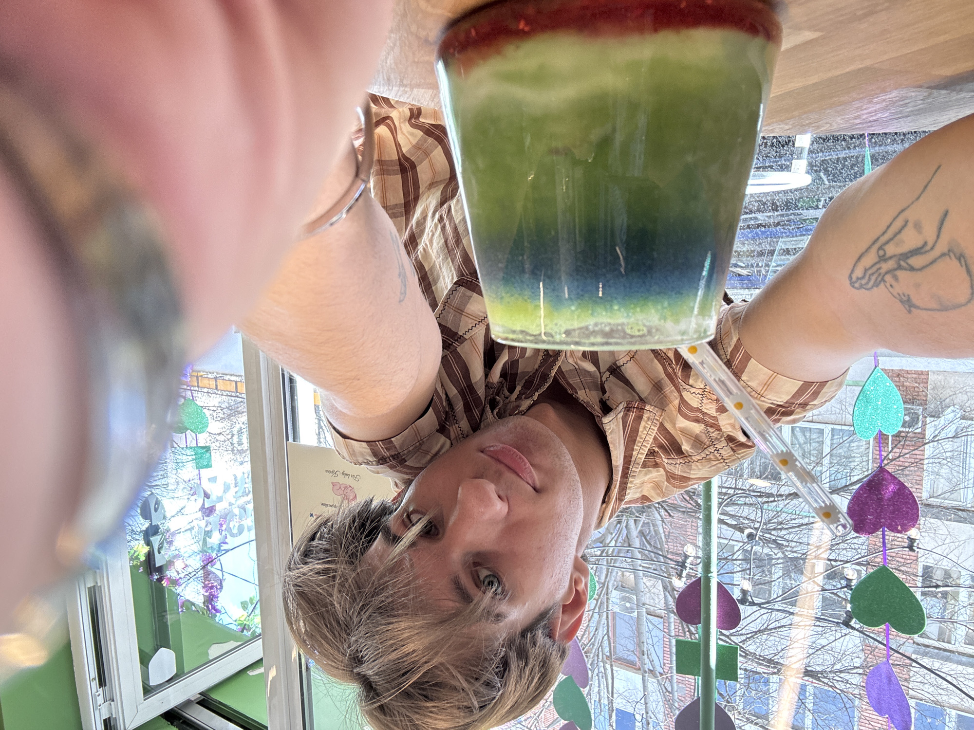 The image displays a close-up selfie of a young, light-haired individual, appearing male, situated inside a cafe or juice bar in Sofia, Bulgaria. The subject wears a brown and tan plaid short-sleeved collared shirt. A tattoo depicting two cupped hands holding a small animal is visible on his left forearm. His left arm is extended, holding the camera, with his face centered in the frame, exhibiting a neutral expression. A silver bracelet is visible on his right wrist.
In the immediate foreground, resting on a light-colored wooden table, is a clear glass containing a layered beverage. The drink features a red bottom layer, a vibrant green middle layer, and a blue-green top layer. A clear plastic straw adorned with small yellow daisy patterns is inserted into the drink.
The background behind the individual features a large window. Suspended in front of this window are iridescent heart-shaped decorations in purple, green, magenta, and teal. A light green interior wall or trim is visible to the right. A sign, partially obscured, displays the text "For baby Karina." Through the window, an urban outdoor scene is visible, characterized by bare tree branches, multi-story buildings with brick and rendered facades, and overhead utility lines, consistent with a street environment in Sofia, Bulgaria.