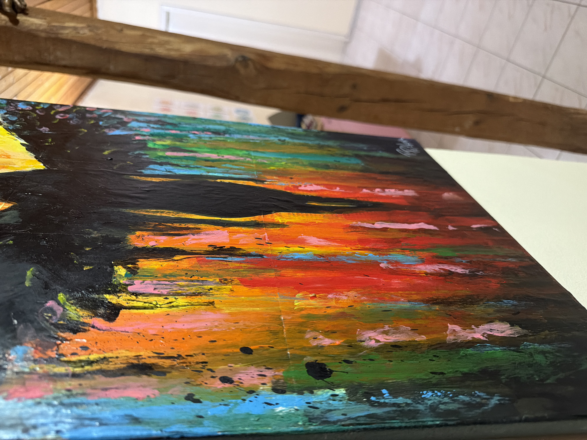 The image displays a close-up perspective of an abstract painting, characterized by vibrant, dripping colors and bold black brushstrokes. The painting is positioned on an easel, leaning against a wooden beam that is part of a rustic interior. The background shows a tiled floor and a wall, hinting at an indoor setting. The painting itself features a dominant black silhouette against a background of vertical streaks of orange, red, yellow, green, blue, and pink. Splatters of black paint are visible throughout the colorful sections. A signature is present in the lower right corner of the painting. There are no people visible in the image. The context provided about Svilengrad, Bulgaria, does not correspond to any visual elements within the image itself.