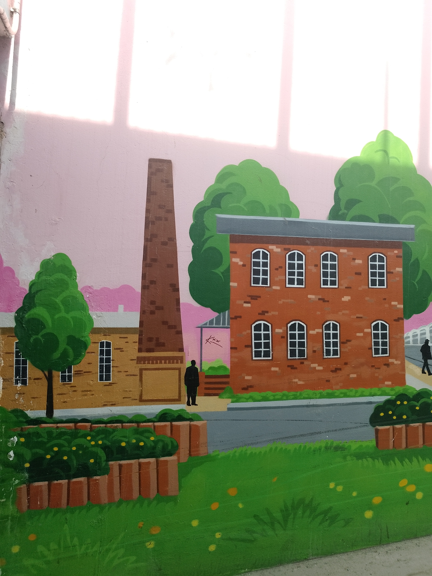 This is a mural depicting an industrial scene, possibly located in the city of Unknown, Unknown. The artwork is painted on a wall and features several buildings: a tan, factory-style structure with windows and a tree in front of it, a tall brick chimney, and a red-brick building with a grey roof and arched windows. Two human figures are included: one standing in front of the chimney and another walking on a pathway behind the red-brick building. The scene includes green trees, bushes, and a lawn with yellow flowers. The background is a light pink color with lighter vertical stripes, possibly representing sunlight or shadows.