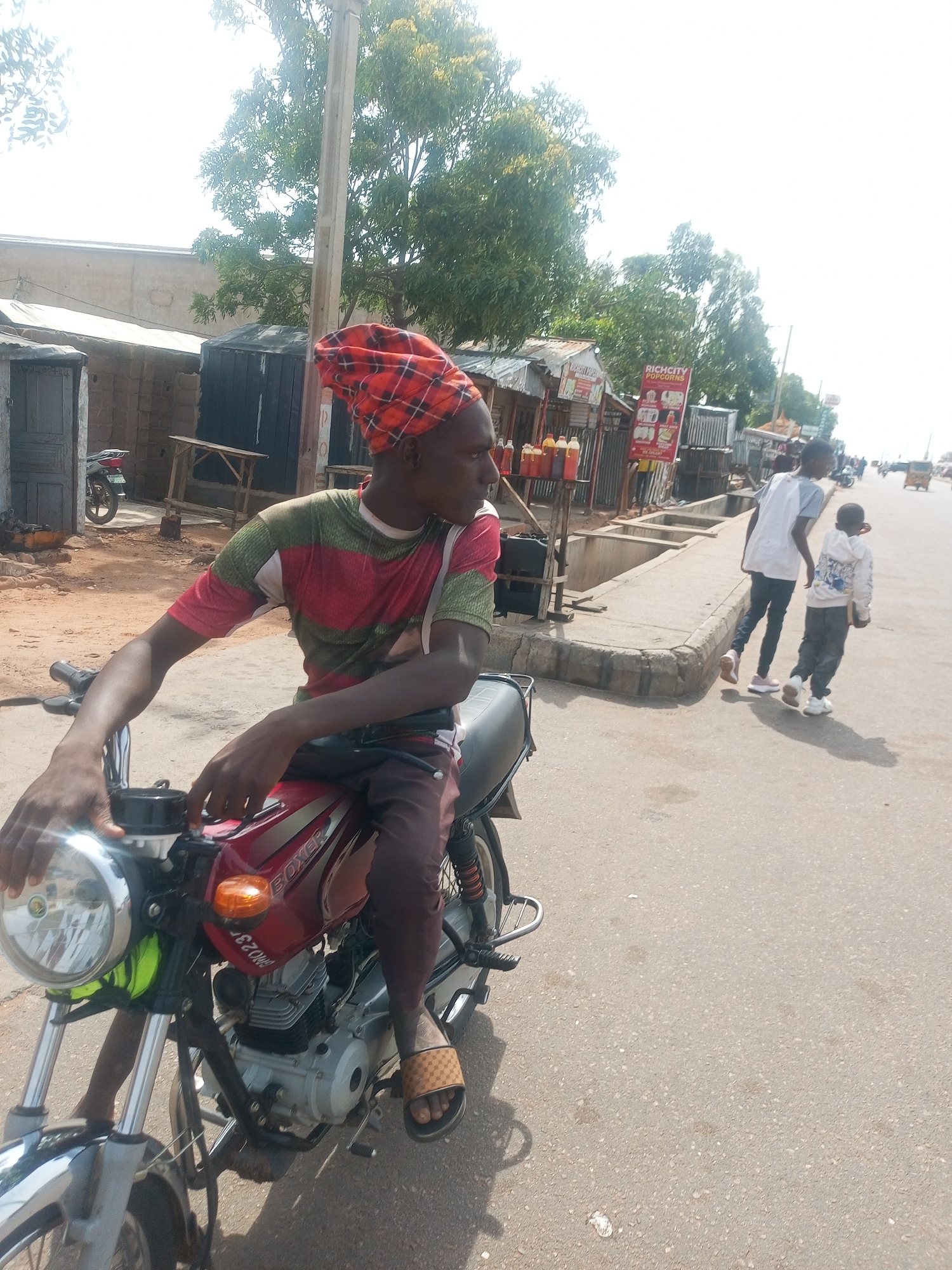 A man wearing a red and black checkered headwrap and a striped t-shirt sits on a red motorcycle in an outdoor urban setting. The motorcycle has the word "BOXER" visible on its side. The man is holding onto the handlebars. In the background, there are small shops with corrugated metal roofs, some wooden structures, and a utility pole. Two other individuals, a taller one in a white shirt and dark pants and a shorter one in a light-colored hooded jacket and dark pants, are walking away from the camera on the right side of the image. The ground is paved, and there are some trees with green foliage visible. The sky is bright and appears to be daytime.