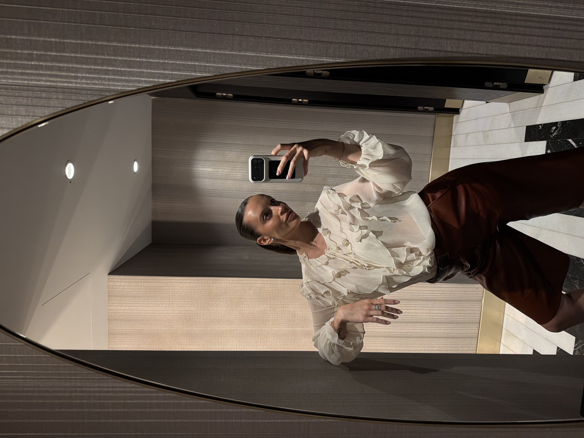 A woman is taking a mirror selfie in a well-lit room with patterned wallpaper. She is leaning against a wall and holding a smartphone in her right hand, angled to capture her reflection. She is wearing a ruffled, cream-colored blouse and brown leather shorts. Her left arm is bent, and her hand rests on her hip. The mirror is oval-shaped with a thin gold frame. The room appears to be a dressing room or a hallway, with a door visible in the background. The floor has a checkered pattern of black and white tiles. The lighting is soft and even, highlighting the textures of the wallpaper and the woman's clothing. The overall scene suggests a moment of personal grooming or preparation. The location is the city of Budva, Montenegro.