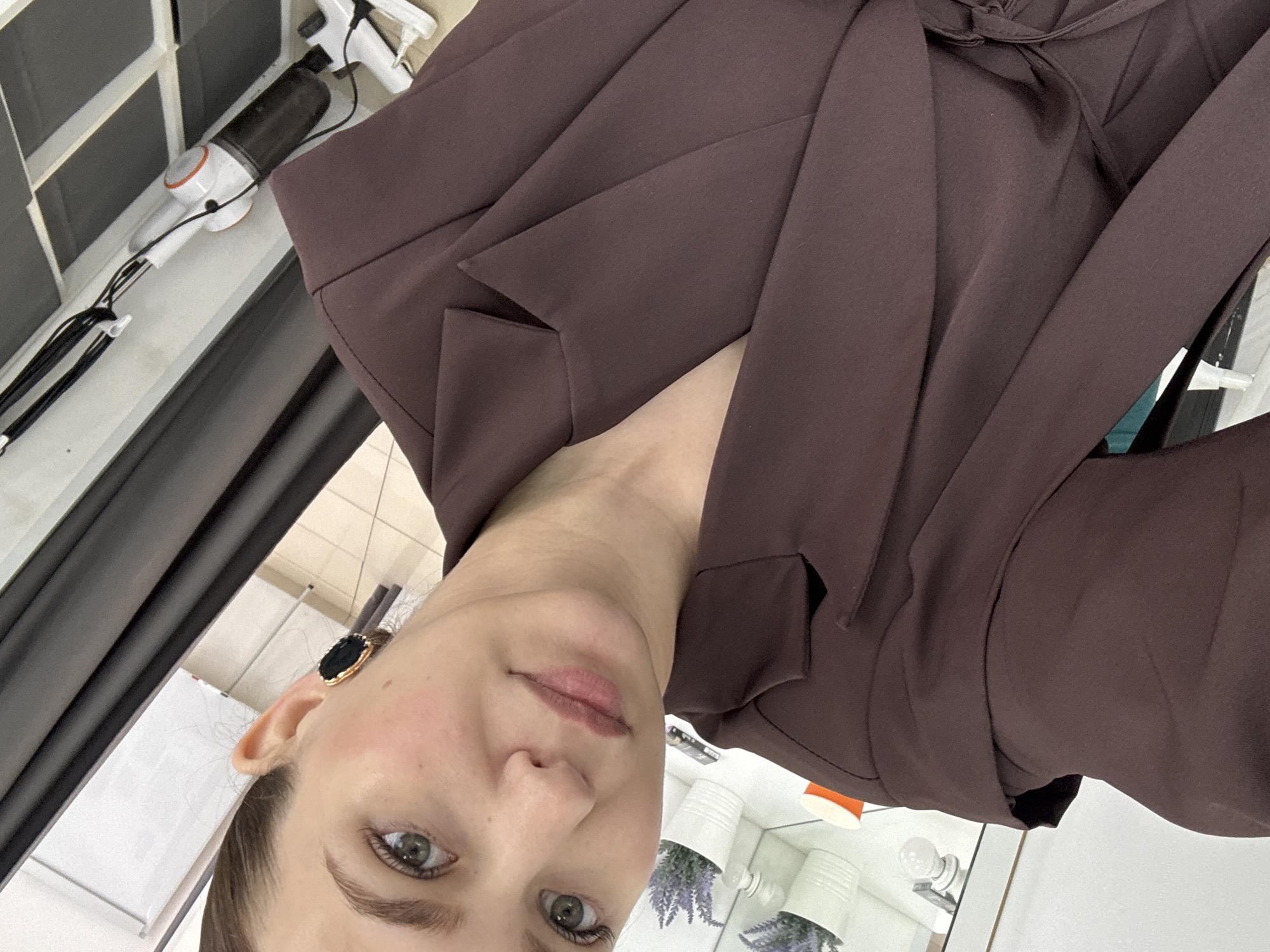 A woman is taking a selfie in what appears to be a studio or dressing room. She is wearing a dark brown blazer. She has on large, dark earrings with gold trim. In the background, there are shelves with plants and lighting fixtures. The overall impression is of a well-lit, tidy indoor space. There is no indication of the city of Chaikovsky, Russia in this image.