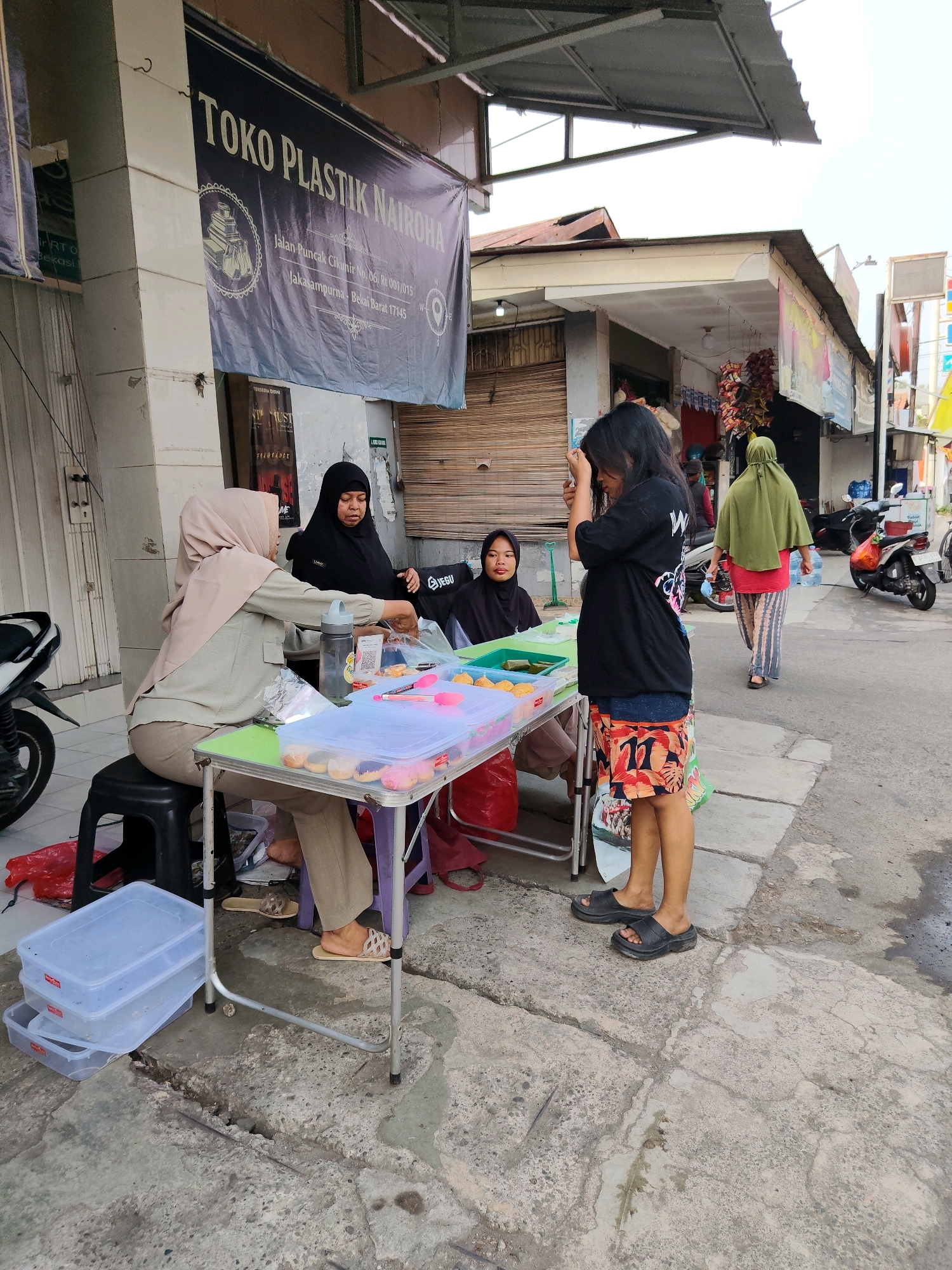 This image captures a street scene in Bekasi, Indonesia, focusing on a small outdoor food stall. In the foreground, three women are present. One woman, wearing a light brown hijab and a long-sleeved shirt, is seated behind a folding table, appearing to arrange items. Another woman, dressed in a black hijab and long-sleeved garment, is standing beside the table, looking downwards. A third woman, the youngest of the group, is standing to the right, facing the stall and touching her hair. She wears a black t-shirt and colorful patterned shorts.
The table is laden with various food items, some in clear plastic containers, including what appear to be donuts or pastries. A water bottle, pink tongs, and plastic bags are also visible on the table. In the background, other individuals are seen walking along the street, and motorcycles are parked. The environment suggests a busy urban area, with buildings and shopfronts lining the street. A banner with Indonesian text, indicating a business named "TOKO PLASTIK NAIROHA," is prominently displayed on one of the buildings. The street itself is paved with concrete, showing signs of wear and tear. The lighting suggests it is daytime, with the sky appearing overcast.