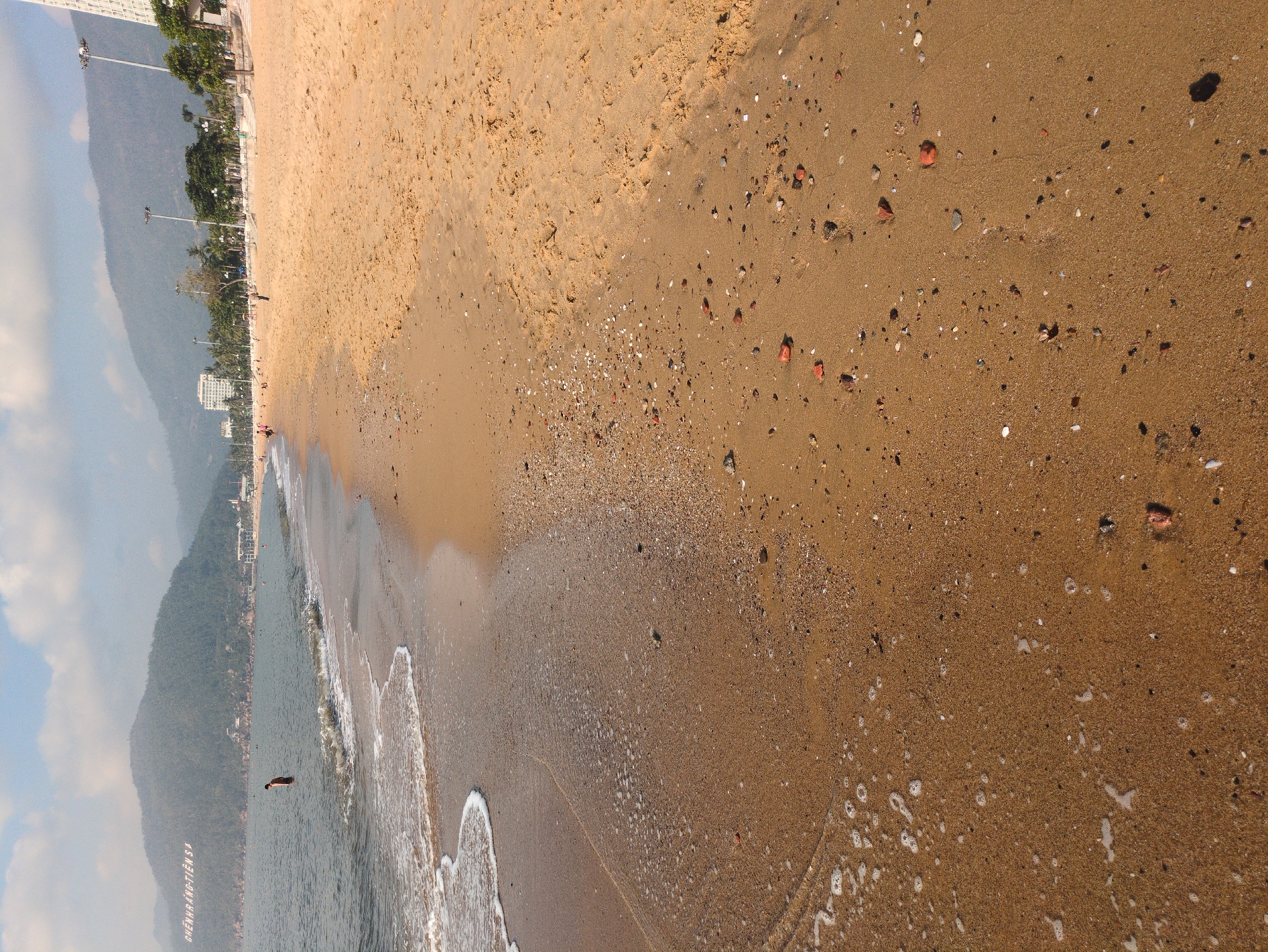 The image depicts a beach in South Quy Nhon Ward, Vietnam. The scene includes a sandy beach with the tide coming in. There is a view of a mountain and some buildings in the distance. There is one person in the water.