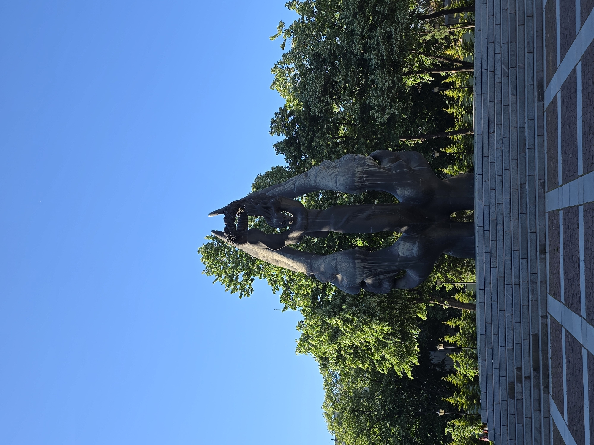 The image shows a metal sculpture of what appears to be multiple figures, possibly abstract in nature, reaching upwards. The sculpture is located outdoors, adjacent to a stone staircase and a patterned brick pathway. Behind the sculpture are trees with dense green foliage. The sky is clear blue. The sculpture appears to be situated in a park or public space in Unknown, Unknown.