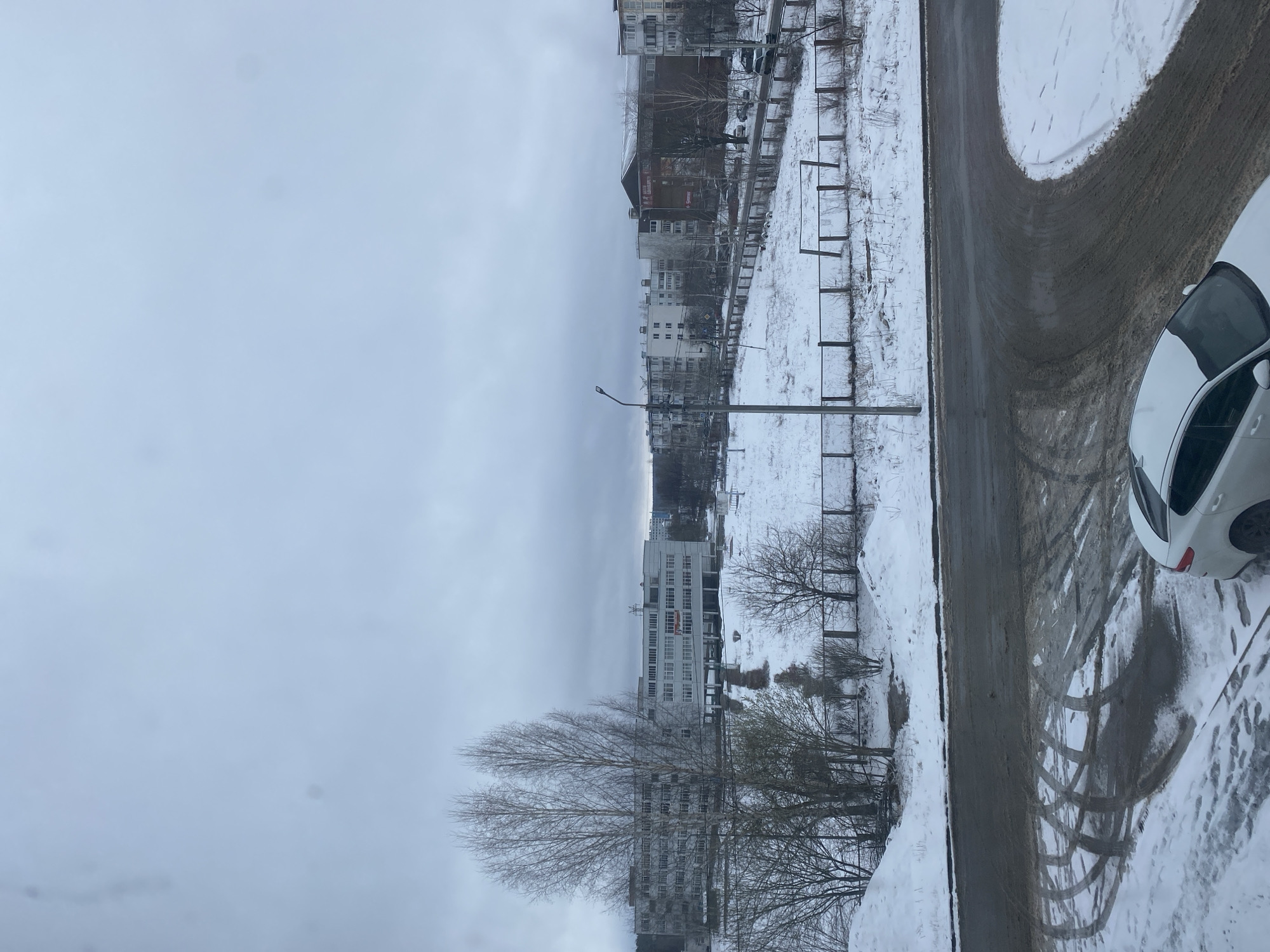 The image shows a winter scene in what appears to be a residential area in the city of Chaikovsky, Russia. The weather is overcast and the ground is covered in snow, with a partially cleared road curving to the right. A white car is parked on the snow-covered ground near the road. In the background, there are several multi-story buildings, typical of urban residential areas in Russia. Fences and utility poles are visible along the edge of the cleared area. Bare trees are interspersed among the buildings and along the roadside. Tire tracks are visible in the snow.