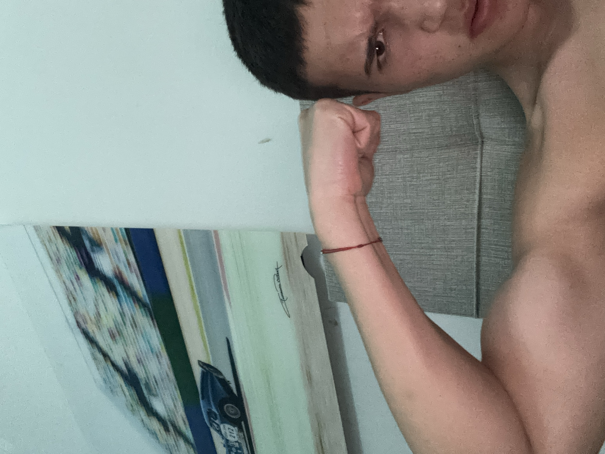 The image shows a person indoors, captured from a tilted, close-up perspective. The subject, with short dark hair, is positioned in the upper right, looking towards the right side of the frame with their head slightly tilted. Their right eye is clearly visible, and their expression is neutral. The person's right arm is bent at the elbow, showcasing a flexed bicep, and a thin red string bracelet is worn on the wrist. Their upper body appears shirtless.
Behind the person's head and shoulder is a grey, textured surface, resembling a pillow or headboard. The background wall is plain white. In the lower left, a decorative board or picture frame leans against the wall. This board features vertical stripes of various colors and includes a partial image of what appears to be a vintage blue race car, with the number "212" visible on its side. An illegible signature is also present on the board.
The activity depicted is the person posing and flexing their arm. The setting appears to be an indoor space, possibly a bedroom. There are no specific visual cues within the image to indicate the stated location of Plovdiv, Bulgaria.