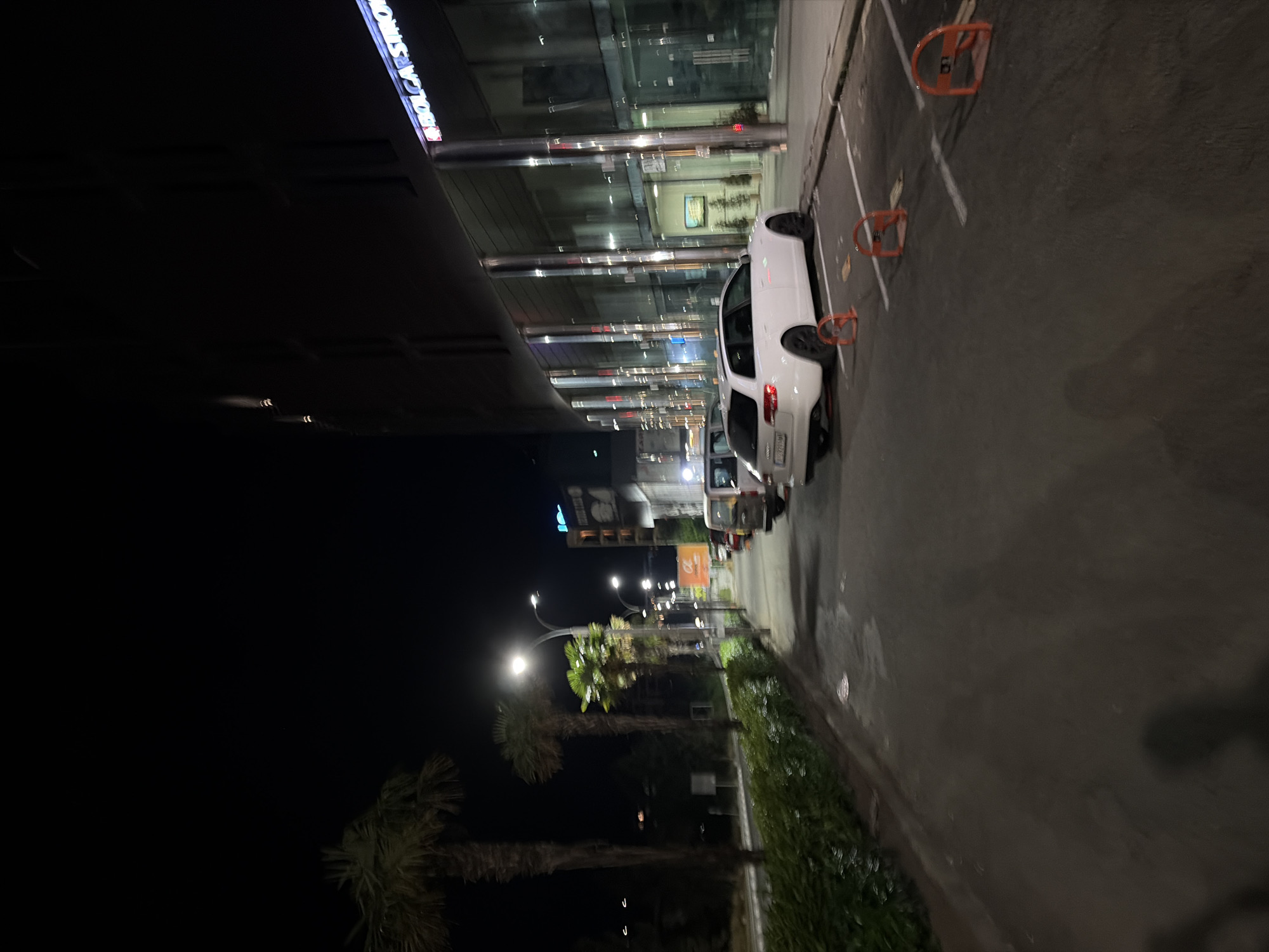 At night, a white Audi station wagon is parked on a street next to a building with a glass facade. The car is in a parking spot marked with orange wheel stops. A palm tree is visible to the left of the car. The building has a sign that says "BOLCAR STRO" in neon letters. There are streetlights illuminating the scene. The street is paved and there is a sidewalk. The overall impression is of a modern, urban environment at night.