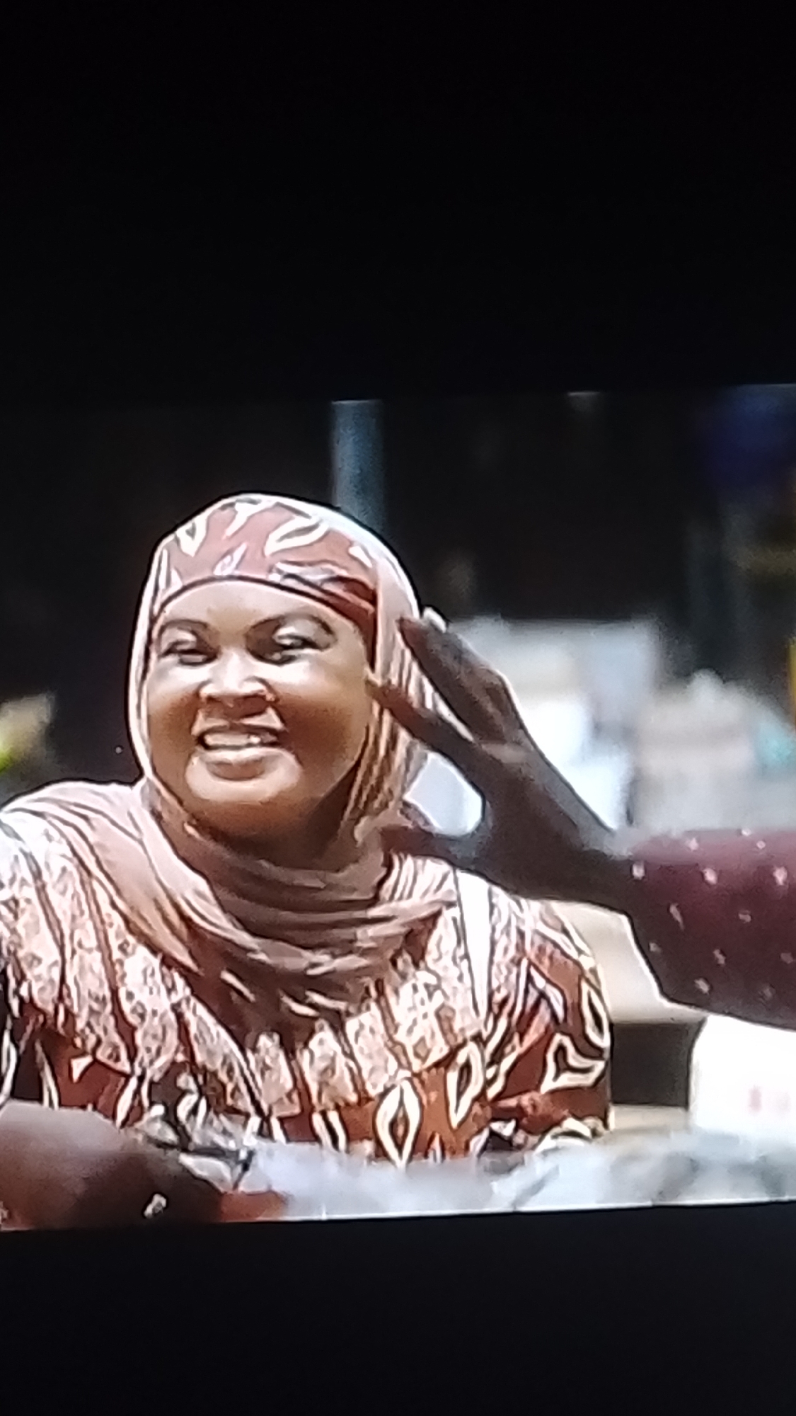 In Lokoja, Nigeria, a woman wearing a headscarf and patterned clothing smiles broadly as another person's hand rests on her head. The woman's face is the primary focus, with her eyes closed and mouth open in a laugh. The background is blurred, but suggests an outdoor setting with some foliage and structures. The woman is also holding something small and white in her hands, possibly food or a small item. The hand on her head appears to be from a person with darker skin, wearing a maroon garment with white polka dots. The interaction suggests a moment of lightheartedness and connection.