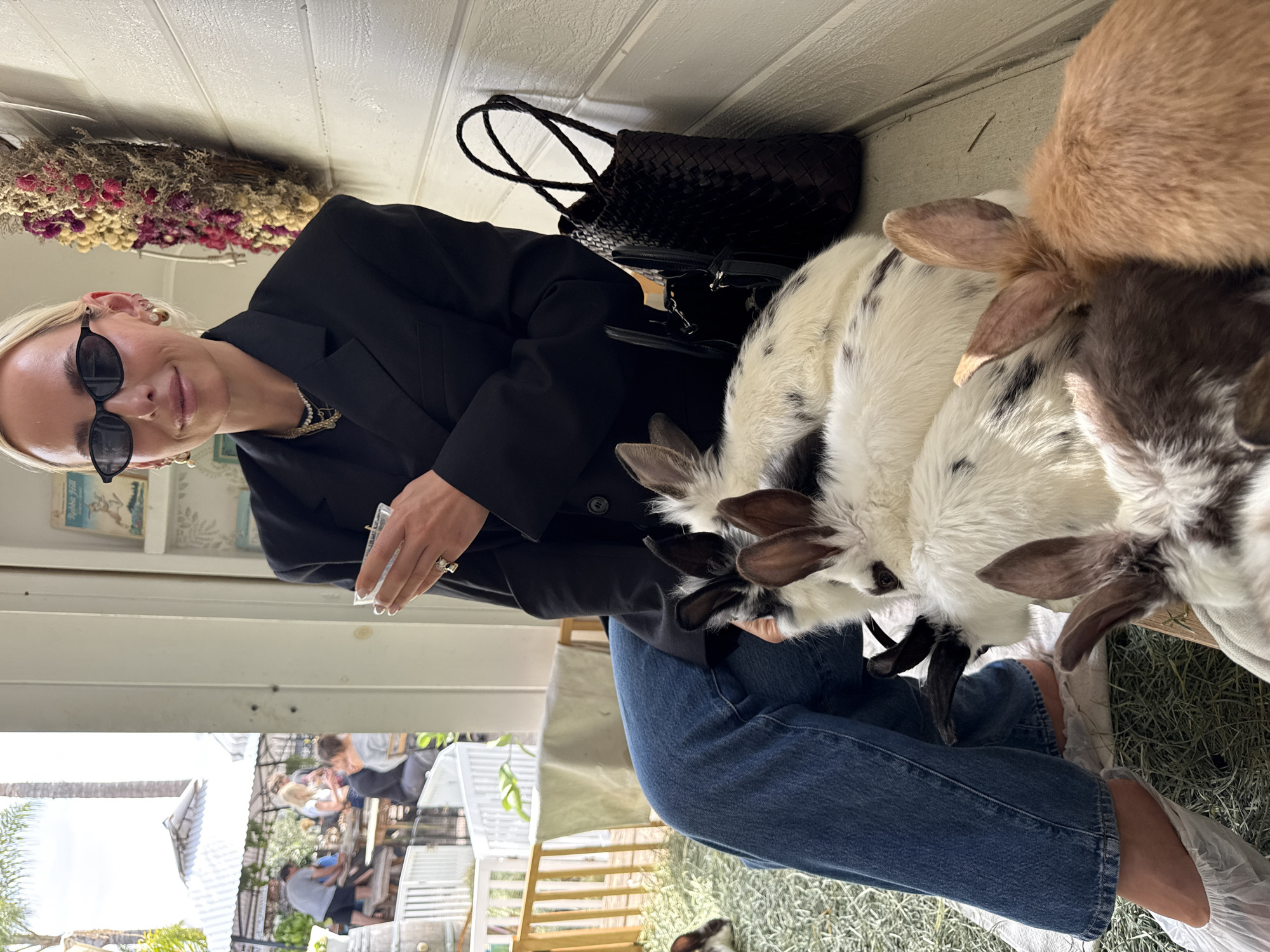 A woman wearing sunglasses and a black blazer is surrounded by several goats that are eating in an enclosure. She is holding a small, clear container in one hand. A black woven tote bag sits near the woman on the ground near the wall and dried flowers are hanging on the wall above. The goats vary in color, including white with black spots, brown, and gray. The woman is wearing blue jeans and transparent plastic shoe covers. In the background through the window of the enclosure, people are seated at tables. The image appears to be taken outdoors. Based on the presence of goats and the exterior appearance, it is likely the setting is the Goat Yoga Solana Beach location in Solana Beach, CA.