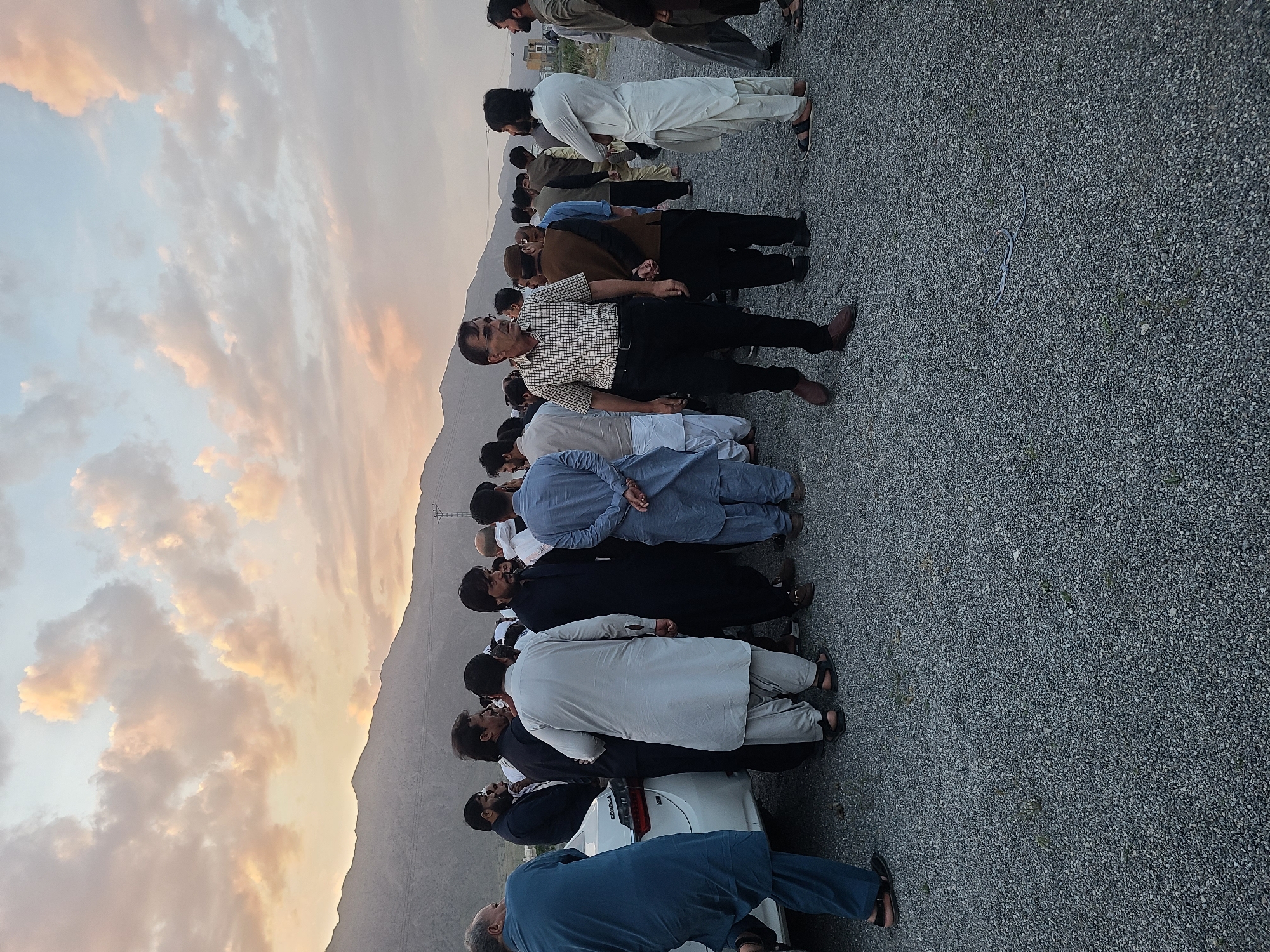 The image depicts an outdoor gathering of a large group of men in Quetta City Tehsil, Pakistan, set against a stunning sky at either dawn or dusk. The sky features soft hues of blue, orange, and pink, with scattered clouds that reflect the gentle, warm light of the rising or setting sun. In the background, a range of mountains or hills stretches across the horizon, characteristic of the region.
The men are primarily dressed in traditional Pakistani attire, including various colors of shalwar kameez, some paired with waistcoats or jackets. A few individuals are also seen in casual Western clothing such as shirts and trousers. They are standing on a gravel-covered ground, arranged in what appears to be rows or a loose formation. Most men are facing away from the camera, with their hands clasped either in front or behind them, suggesting an organized assembly. Their postures and expressions convey a sense of attentiveness or solemnity, indicating a formal event or observance, possibly a communal prayer or a community meeting.
In the foreground and mid-ground, some individuals are looking towards the camera, while others maintain their gaze in the direction of the main group. A white Toyota Corolla car is partially visible on the far left side of the frame, with the word "COROLLA" clearly identifiable on its rear. A blue string or rope lies on the gravel ground in the mid-ground. The overall scene captures a moment of collective gathering in a natural, open setting, highlighting both the community and the picturesque landscape.