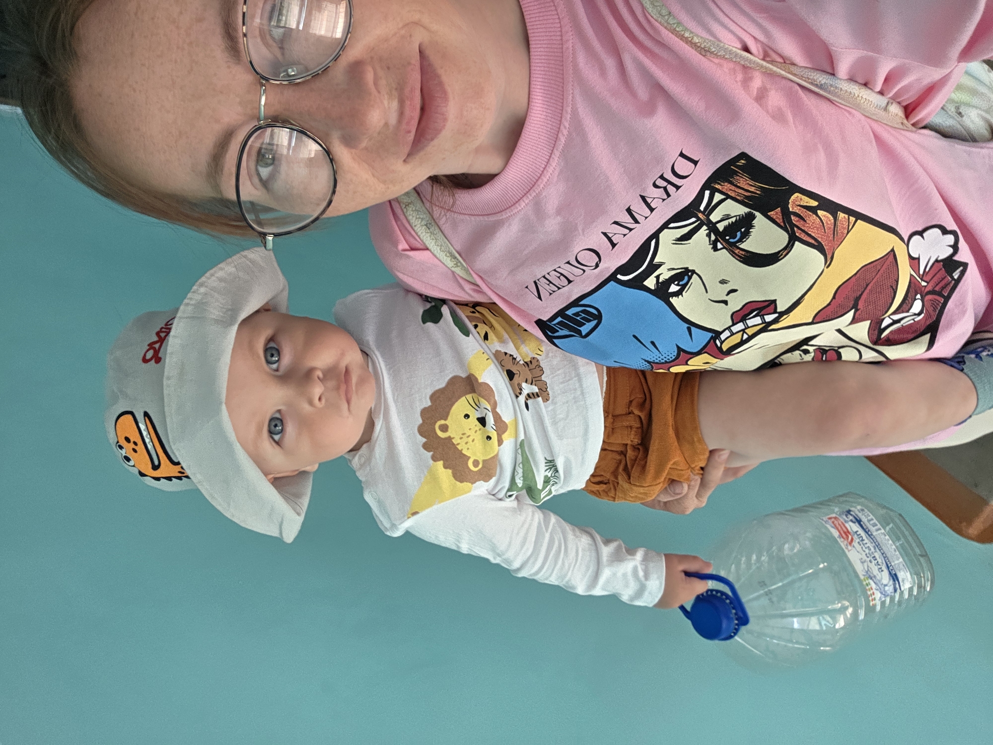 A selfie shot depicts a light-skinned woman wearing round-rimmed glasses and a pink t-shirt with a "Dream Queen" comic book style graphic, positioned above a baby with striking blue eyes. The baby, dressed in a white, long-sleeved shirt adorned with cartoon lions and other animals, along with brownish shorts, holds a large, empty plastic water bottle with a blue handle. The baby also wears a light-colored hat with a cartoon dinosaur. The background is a solid light blue. The image was taken in the city of Chaikovsky, Russia, based on the Cyrillic text on the water bottle label.