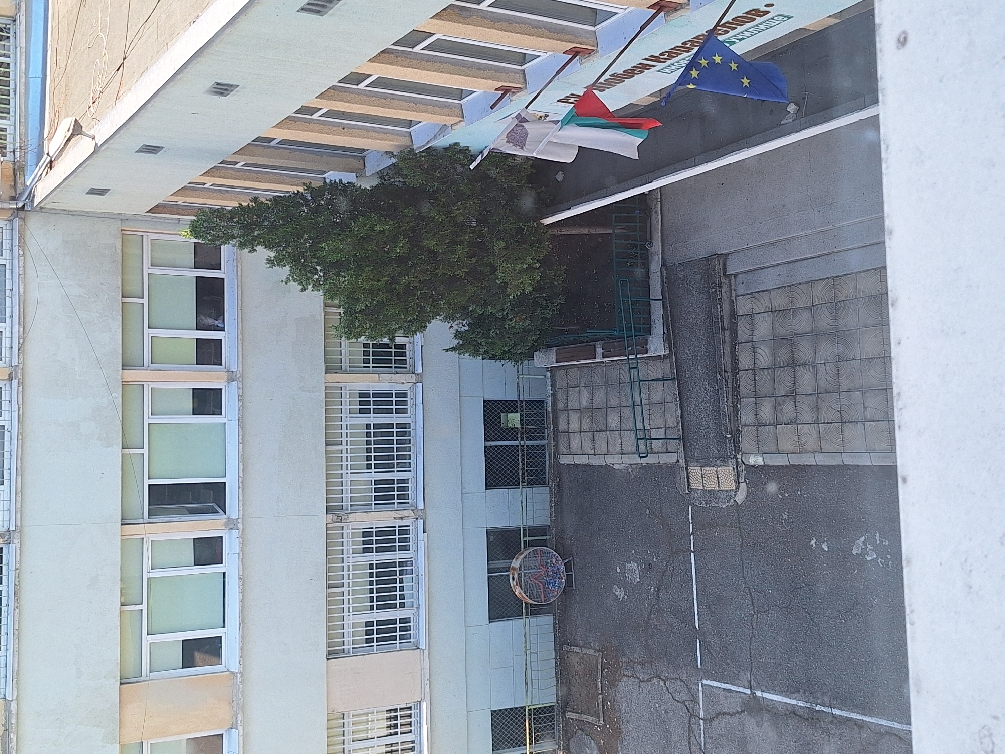 The image captures a view from above, looking down onto the exterior of a light-colored, multi-story building, possibly a school or institution, located in Varna, Bulgaria. The architecture suggests a post-communist era design with long rows of windows, many of which have metal bars. The building has a flat, tarred roof.
In the foreground, a portion of a white railing or windowsill is visible, indicating the vantage point is from another elevated structure. Below, a courtyard area is seen. This courtyard is paved with asphalt and features a large, circular, metal bin with a distinctive pattern, containing what appears to be trash or recycled materials. A fence separates this area from a lower section of the building.
To the right of the building, a tall, conical evergreen tree stands prominently. Adjacent to the tree, a wall displays several flags. A Bulgarian flag, white, green, and red, is draped. Below it, a European Union flag, blue with a circle of yellow stars, is affixed to the wall. A banner with Bulgarian text is also partially visible on the building's exterior. A white cloth is hanging near the flags.
The overall scene is one of an urban environment, likely a schoolyard or institutional grounds, under daylight conditions. The presence of flags suggests a patriotic or official event.