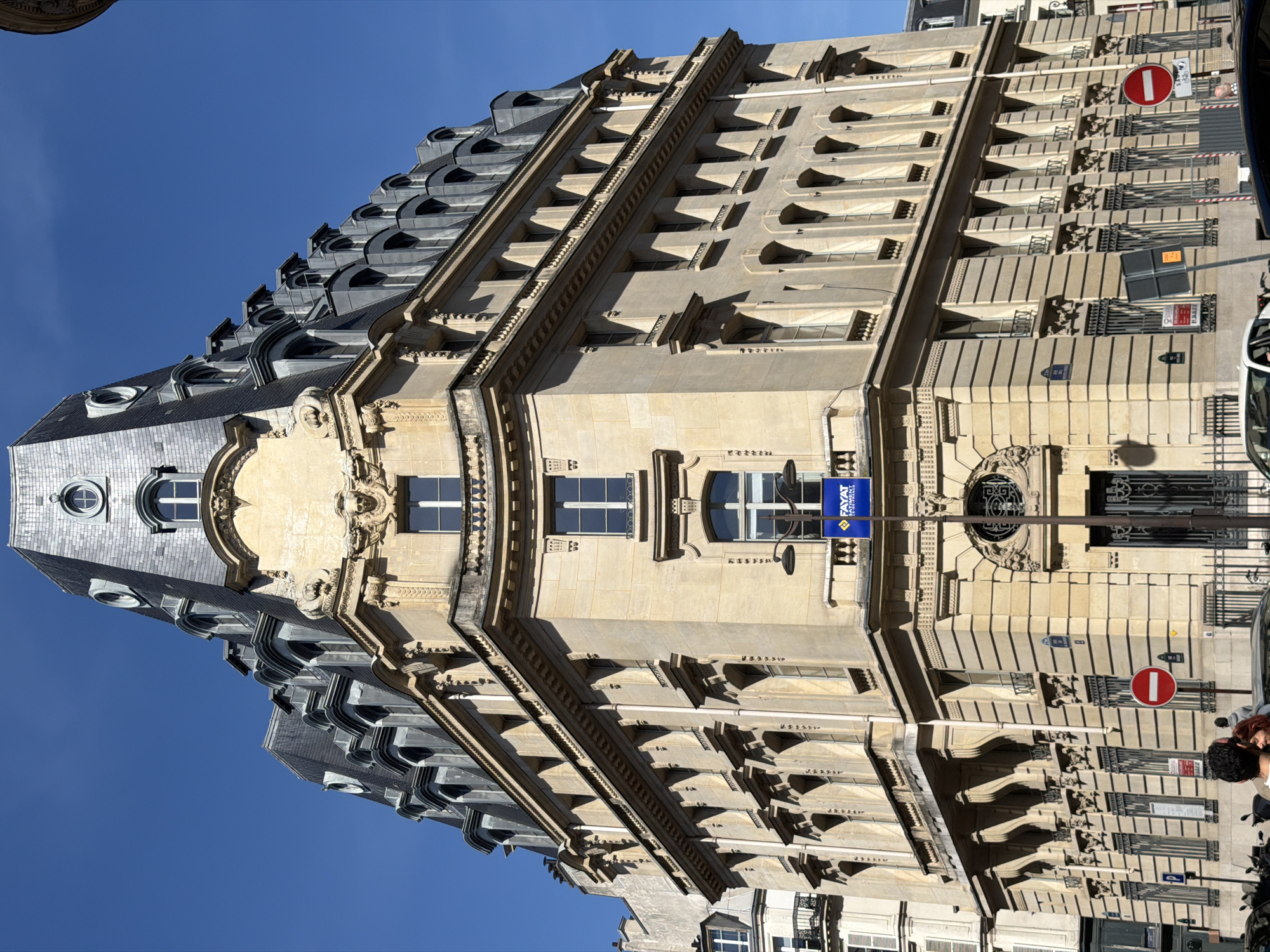 The image depicts a multi-story building in Paris, France, exhibiting classic Parisian architecture with ornate facade details and a steeply pitched mansard roof punctuated by rounded dormer windows. The building's exterior is predominantly constructed from light-colored stone, likely limestone, with decorative moldings, window frames, and cornices. A commercial sign, partially visible, suggests the presence of a business on the ground floor. A "Do Not Enter" sign is affixed to the building near a metal railing. People, vehicles, and signs of street activity can be seen in the foreground, indicating an urban environment. The sky is clear and blue. The building's location appears to be on a street where there is a combination of residential and commercial buildings.