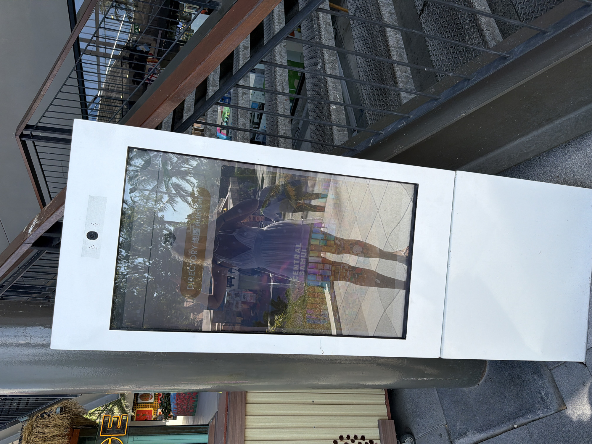 A full-body reflection of a person wearing a light-colored dress is visible in a large, rectangular digital directory kiosk. The kiosk is positioned outdoors, adjacent to a metal staircase. The directory screen displays text that appears to say "DIRECTORY" in English and possibly Thai characters, as well as "CENTRAL SAMUI" which indicates the location as Central Samui in Ko Samui District, Thailand. The person is standing, facing the kiosk, with their arms raised slightly as if to take a selfie. The reflection shows the person's bare legs and feet, and the dress is sheer, revealing a colorful patterned garment underneath. In the background, palm trees and a street scene are visible through the reflection. The kiosk itself is white with a black frame around the screen. To the left of the kiosk, a grey cylindrical pillar and a portion of a building with yellow and green accents are visible. To the right, the metal staircase ascends, with dark grey treads and black metal railings. The ground is paved with grey tiles.