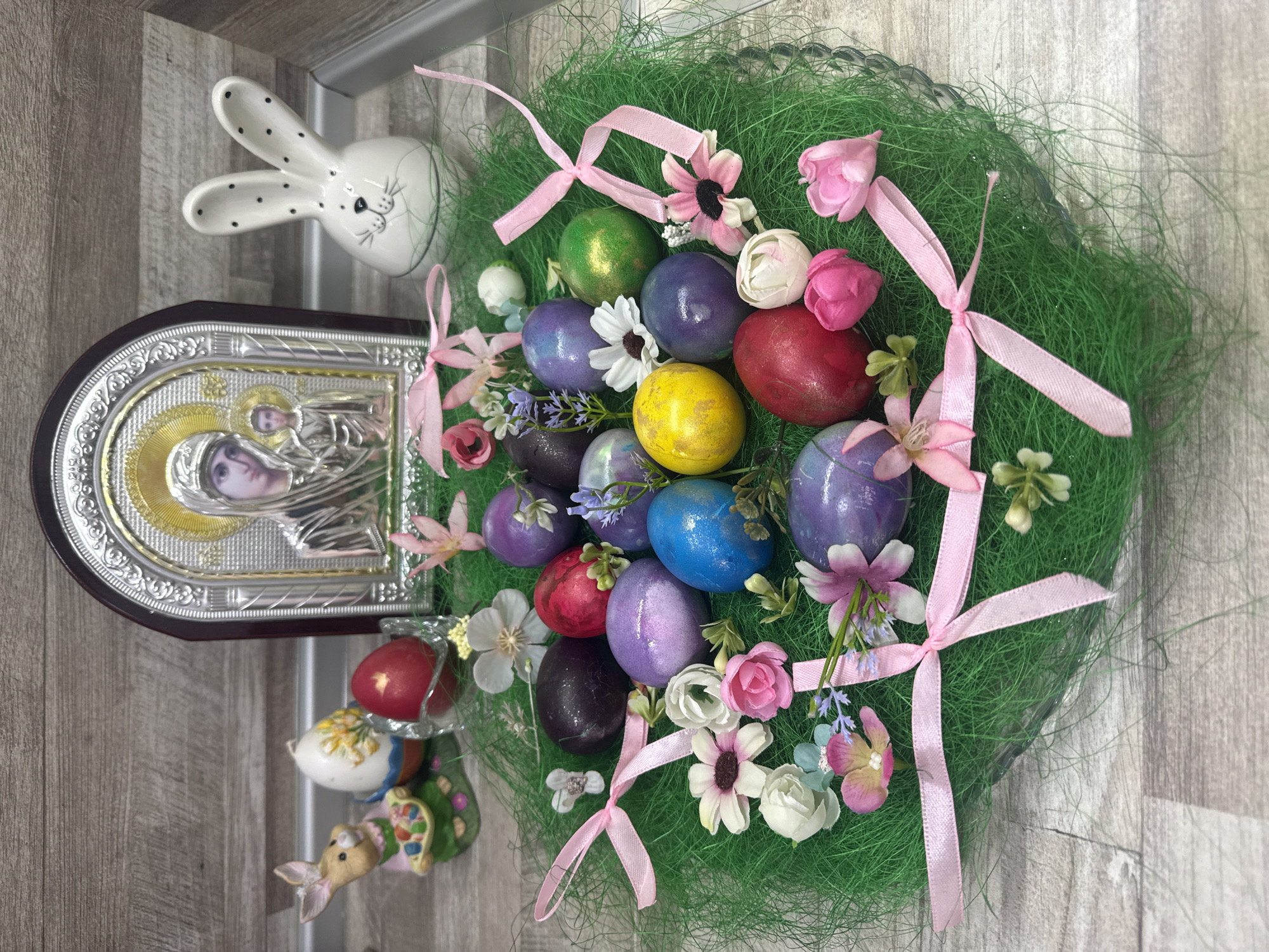 The image shows a still life arrangement on a wood-grain surface, seemingly indoors. Dominating the scene is a green, circular Easter wreath, adorned with colorful Easter eggs in hues of gold, green, purple, red, and blue, interspersed with artificial flowers in shades of pink and white, tied with pink ribbons. To the left of the wreath, there is a religious icon with silver detailing and an image of a woman with a child. Next to the icon, there's a white ceramic bunny head with black polka dots and a ceramic bunny figurine with colorful patterns. The scene suggests Easter decorations, potentially related to religious practices. Varna, Bulgaria is not identifiable.