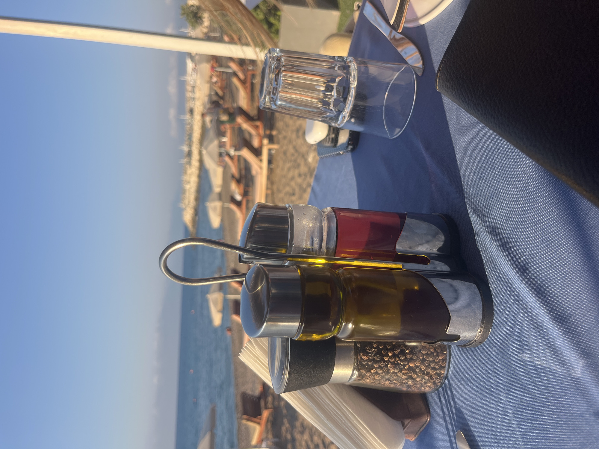 The image shows a close-up view of a table set for a meal at a beachside restaurant in Polis Chrysochous, Cyprus. The table is covered with a blue tablecloth, and in the foreground, there is a set of salt and pepper shakers, one containing whole peppercorns and another holding olive oil. To the right, there is a clear glass that is inverted, and a knife with a white handle rests on a white plate. In the background, the beach and the sea are visible, with lounge chairs and umbrellas suggesting a vacation setting. The sky is clear and blue, indicating a sunny day. No people are directly in focus or interacting in the image.