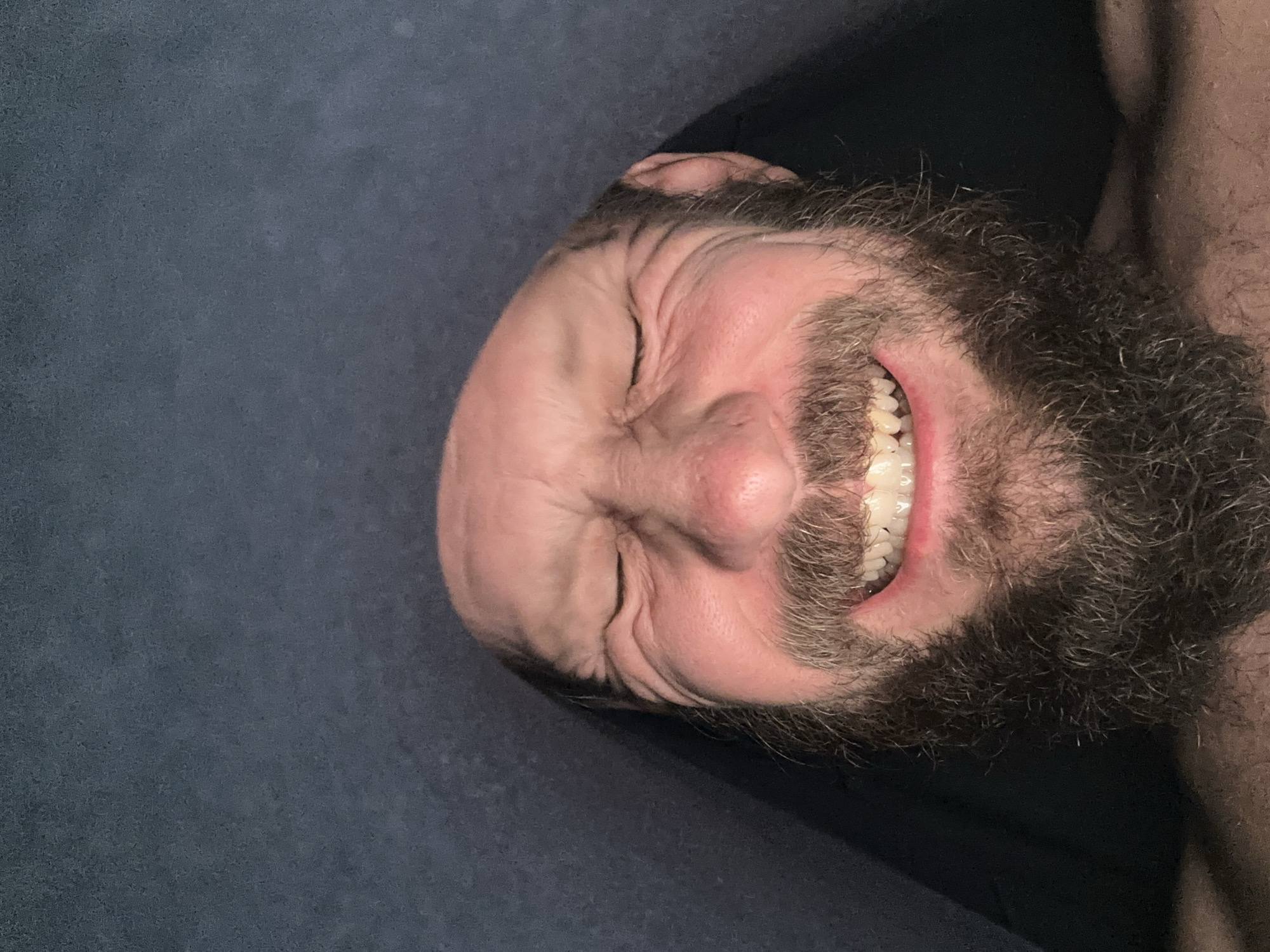 A close-up, upside-down image shows a person with a bald head, closed eyes, and a dense, grey-streaked beard grinning, exposing white teeth. The person's skin is fair and wrinkled, and the background appears to be a dark, solid-colored surface. The scene is not identifiable as being in Fethiye, Turkey, or any specific location.