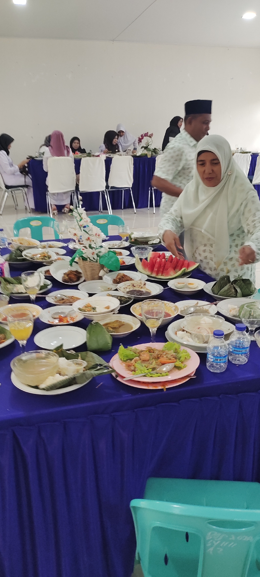 In Meucat, Indonesia, a gathering is underway, likely a celebratory meal given the abundance of food and the presence of multiple people. A long table is laden with a variety of dishes, including what appears to be rice wrapped in leaves, cooked meats, a large platter of sliced watermelon, and bowls of soup or curry. Several individuals, mostly women wearing hijabs, are seated or standing around the table, interacting with the food and each other. One woman in a light-colored hijab is actively arranging slices of watermelon on a plate. The room has white walls and a muted lighting, suggesting an indoor event. The table is covered with a vibrant blue tablecloth, and chairs with white coverings are visible in the background. A floral arrangement sits in the center of the table, adding a decorative touch. The overall scene conveys a sense of communal dining and festivity.