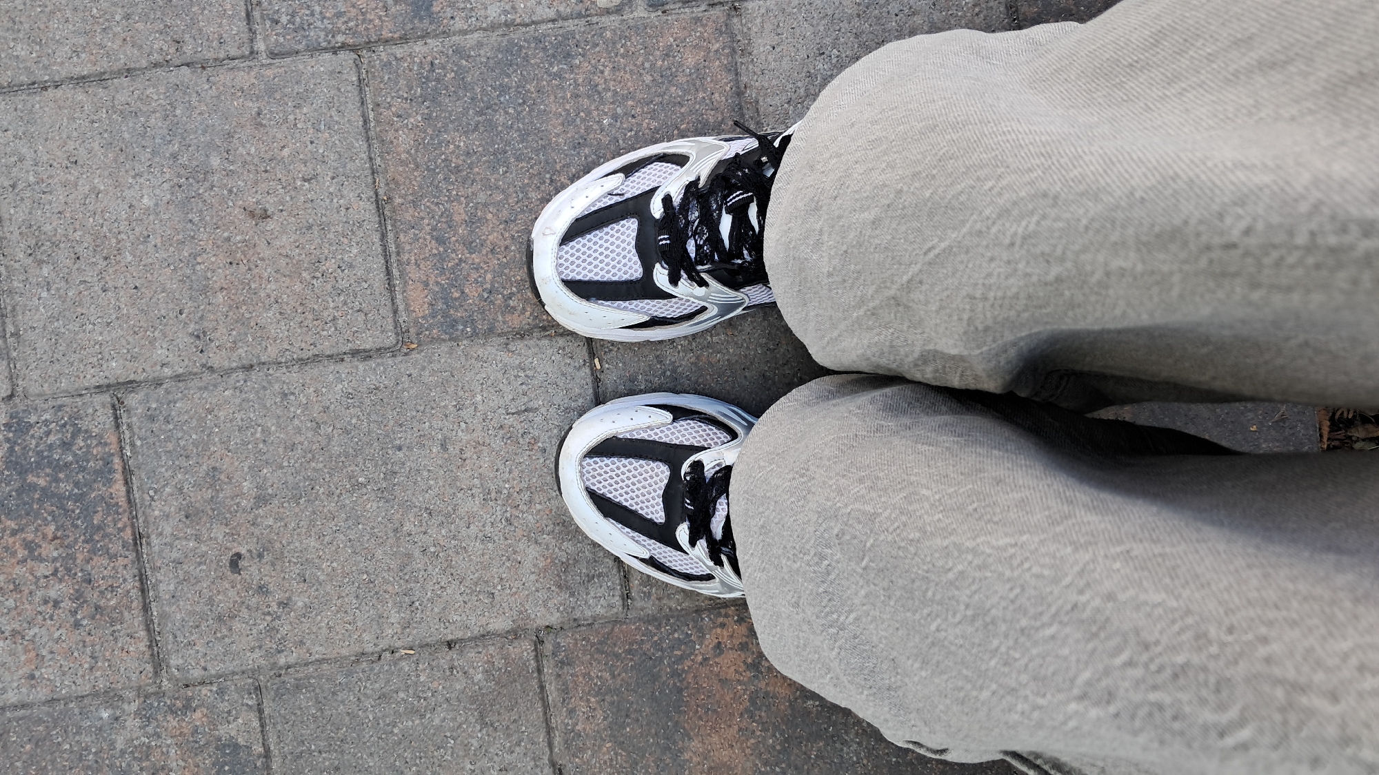The image shows a top-down view of a person's legs and feet wearing light gray pants and white and black athletic shoes. The person is standing on a paved surface made of rectangular gray and reddish-brown bricks. The shoes have white mesh uppers with black accents and laces. The pants are loose-fitting and appear to be made of a textured fabric. The setting suggests an outdoor environment, possibly a sidewalk or a plaza. There is no visible text in the image. The lighting indicates it is daytime. The ground appears slightly damp in places. The overall mood is casual and everyday. The location context is Ternopil, Ukraine.